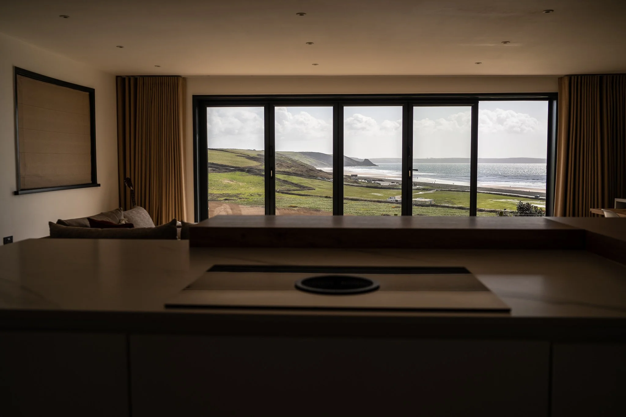Living room with large window overlooking green fields, cliffs, and ocean, with curtains and a sofa visible.