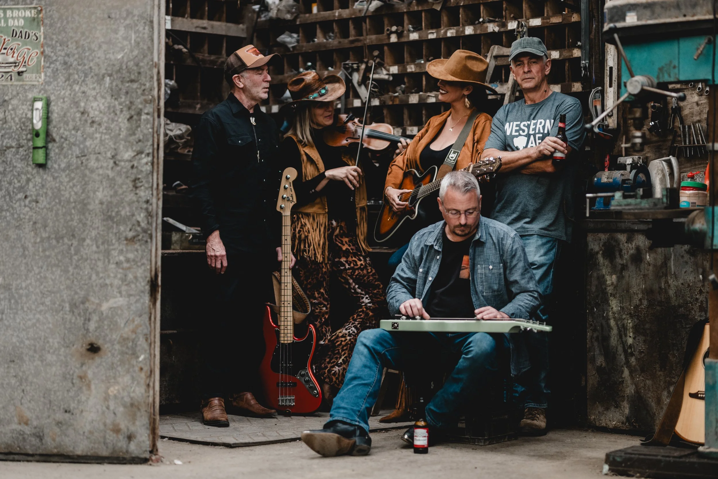 A group of six musicians in a cluttered workshop, with some playing instruments and others preparing to perform. The group includes a woman with a violin, a woman playing an acoustic guitar, a man sitting and adjusting a pedal or effects board, and o