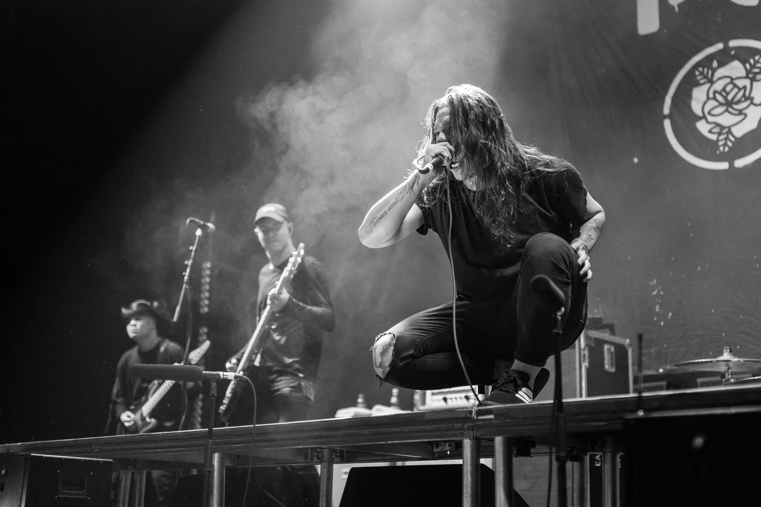 A black and white photo of a band performing live on stage. The lead singer, with long hair, is crouched on a raised platform, holding a microphone close to her face, and appears to be singing or shouting passionately. Polari band