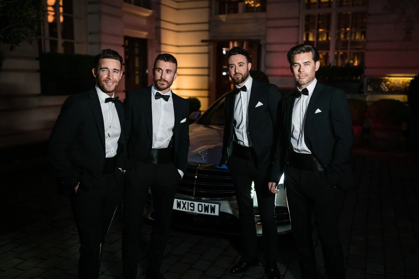 Four men in tuxedos standing in front of a luxury car, dressed for a formal event at night. The OVERTONES music group.