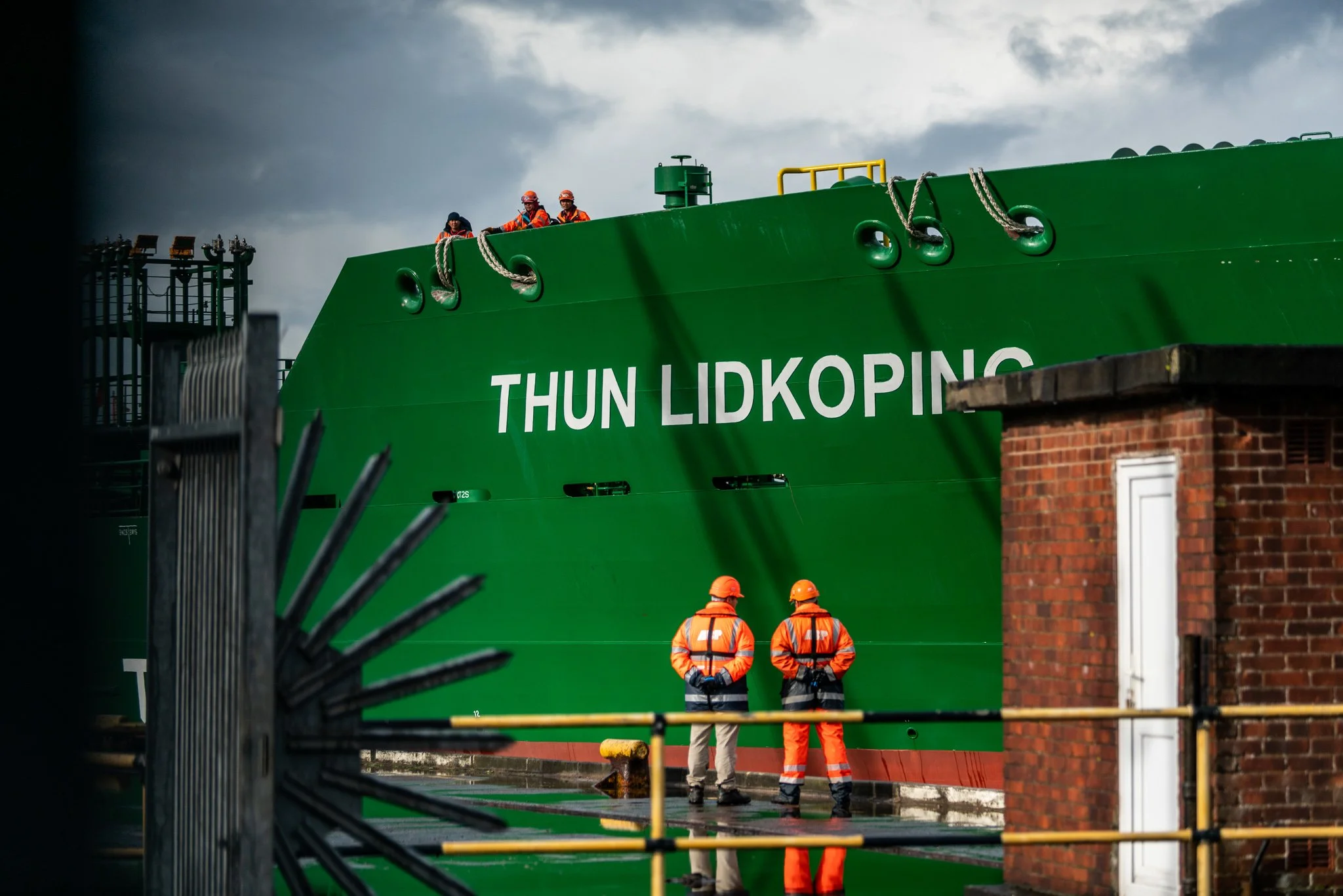 A large green ship named 'Thun Lidkoping' is docked at a port with two workers in orange safety gear standing on the dock and three workers on top of the ship.