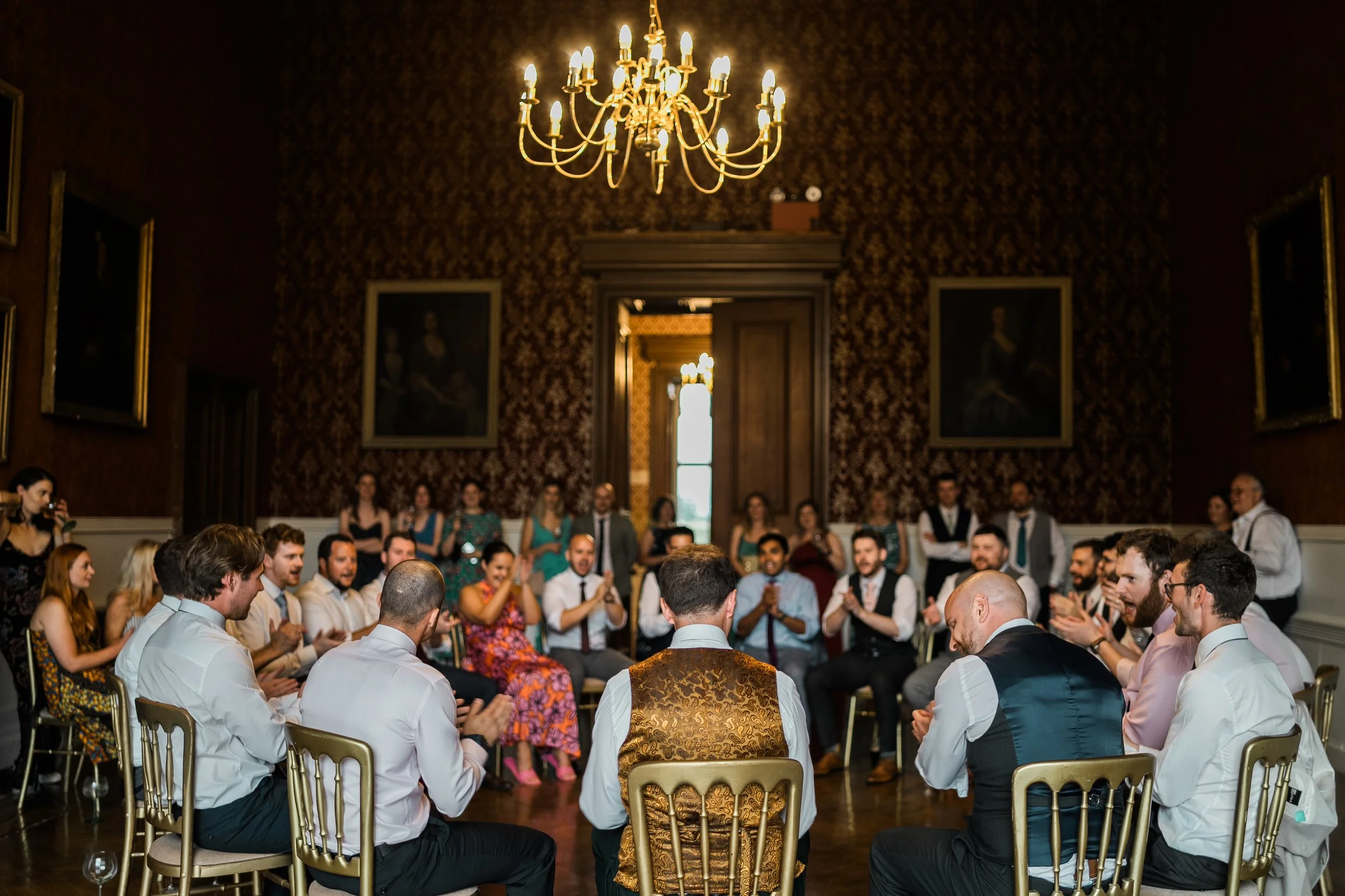 People gathered in a circle in an elegant, vintage-style room with a chandelier, textured wallpaper, and large paintings, celebrating a wedding or special event.