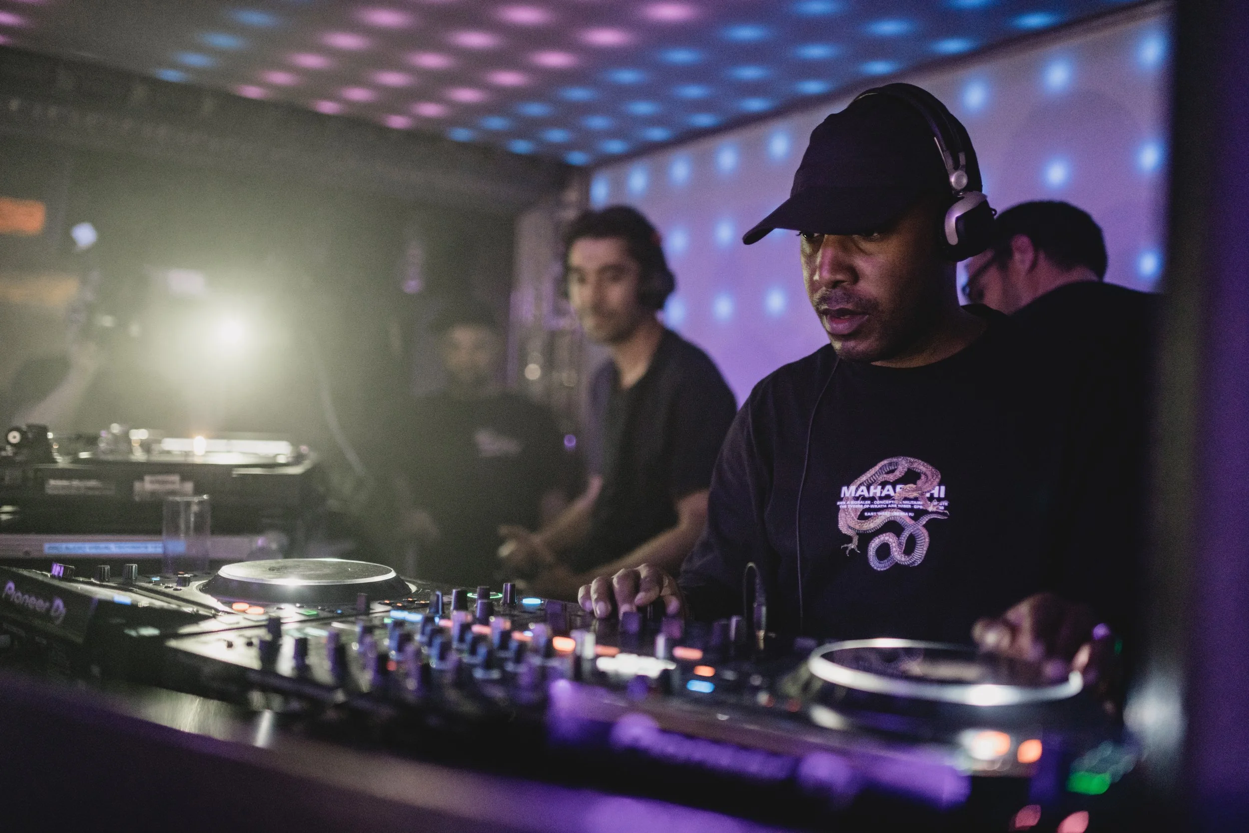 A DJ wearing a black baseball cap and headphones operates DJ equipment in a dimly lit club with colorful LED lights. Shy Fx DJ Photography. Promotional Music Photography.