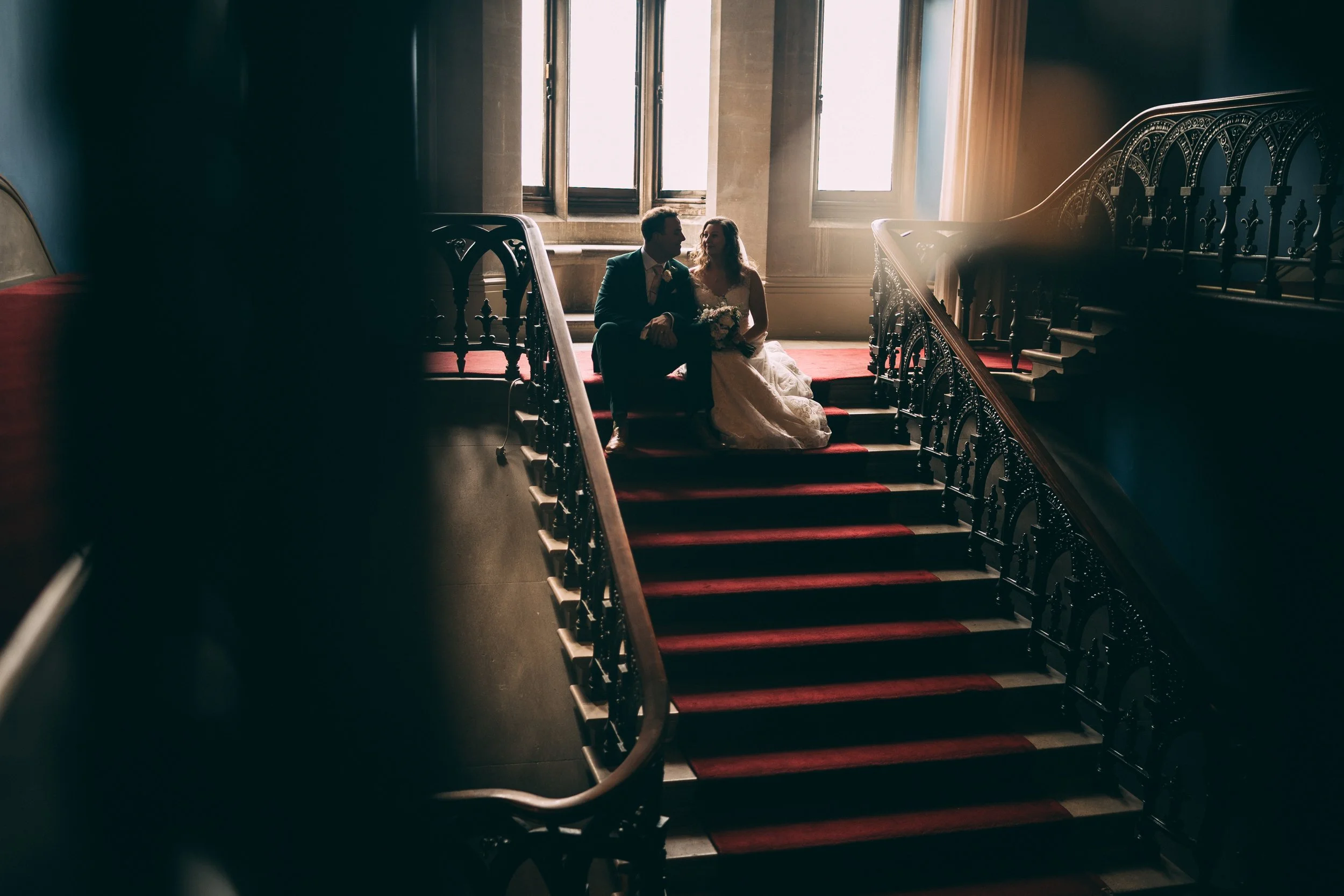 Couple sitting on a staircase, with sunlight coming through large windows behind them, in a historic building with ornate railing and dark walls.