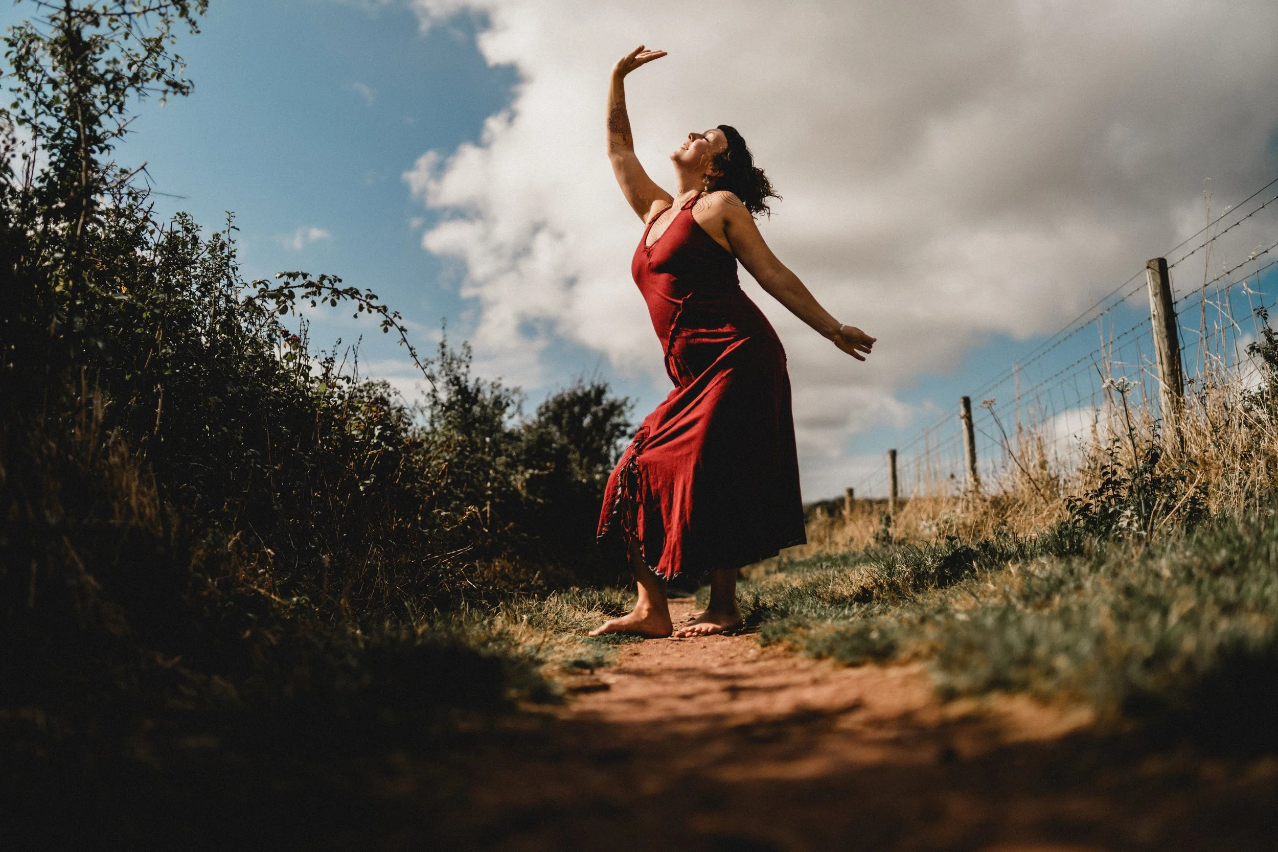 A woman wearing a red dress dancing barefoot on a dirt path outdoors, with a barbed wire fence and bushy vegetation in the background, under a cloudy sky.