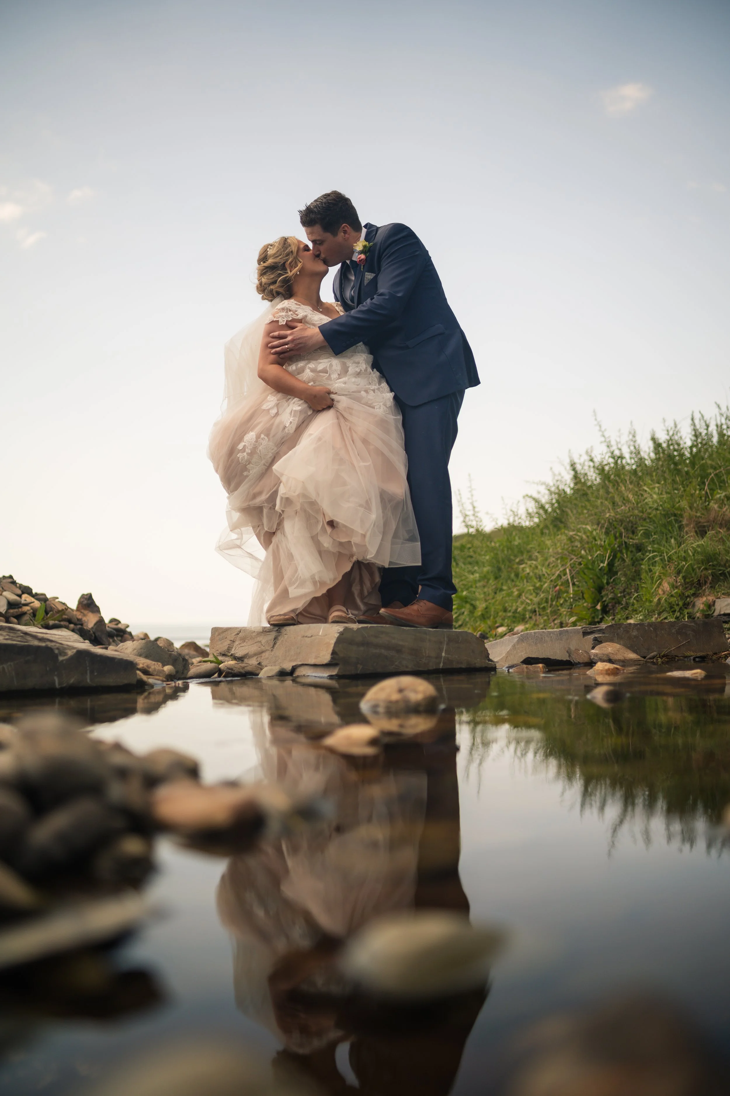 A bride and groom kissing outdoors on rocks near water, with a reflection in the water and a clear sky in the background.