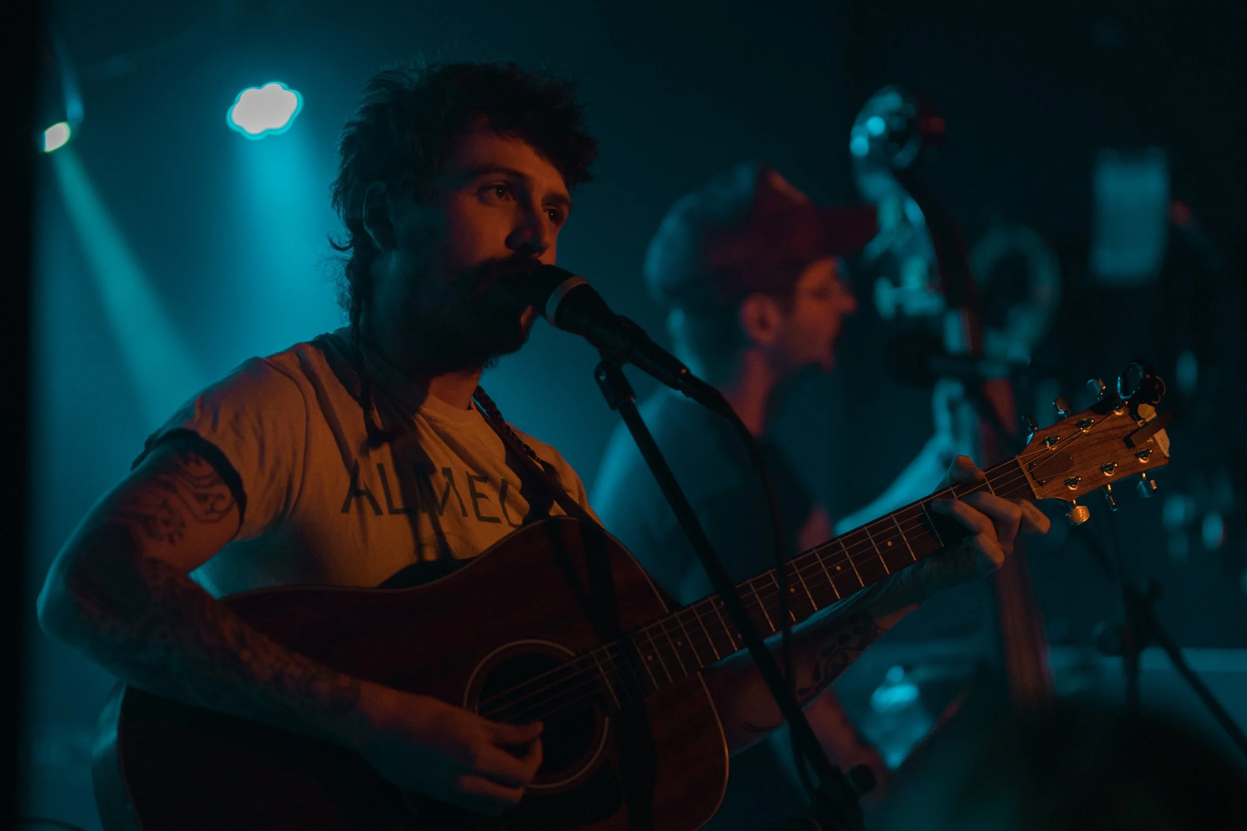 A young man with tattoos on his arms singing into a microphone while playing an acoustic guitar on stage, with another musician in the background, illuminated by blue stage lights. Nick Shoulders country artist.