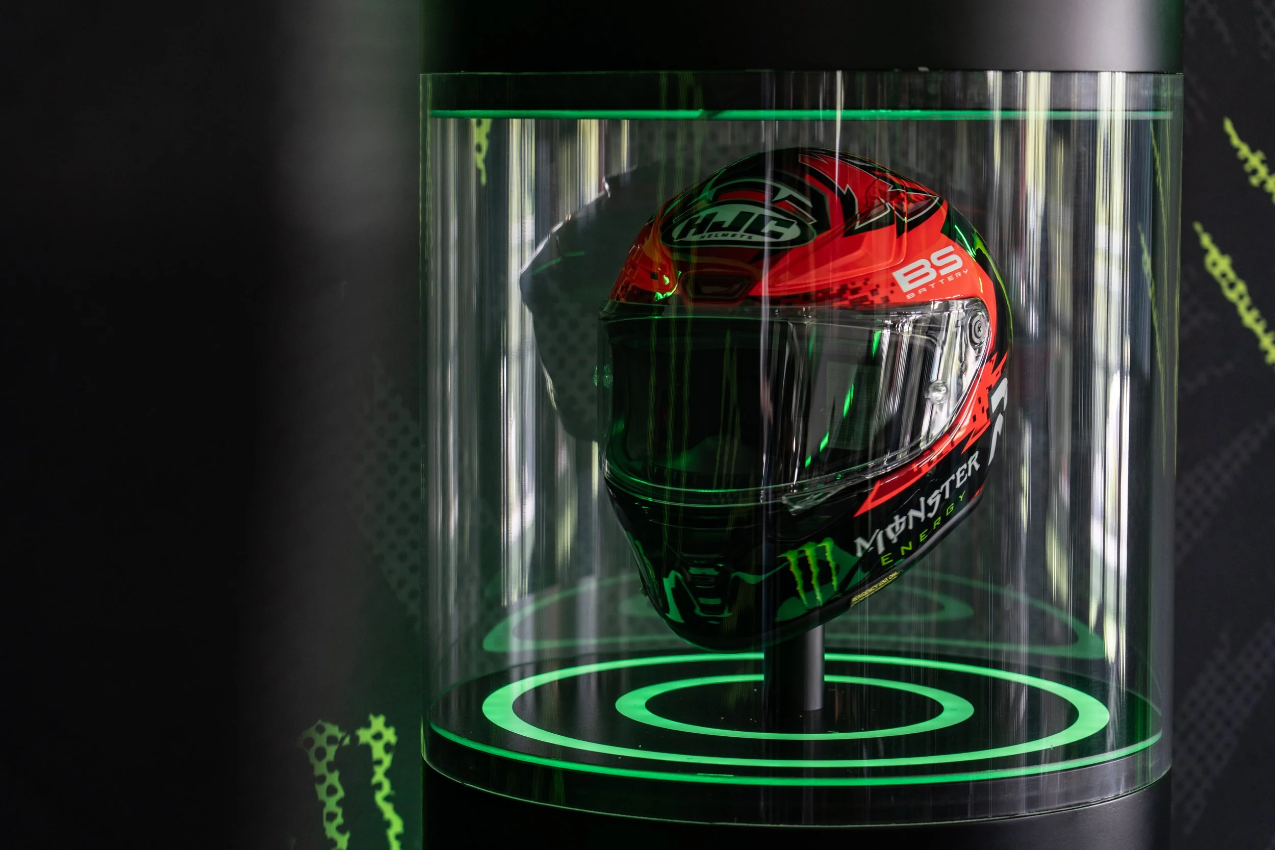 A racing motorcycle helmet in a transparent display case with green lighting, featuring red, black, and green colors, and various sponsor logos including Monster Energy.