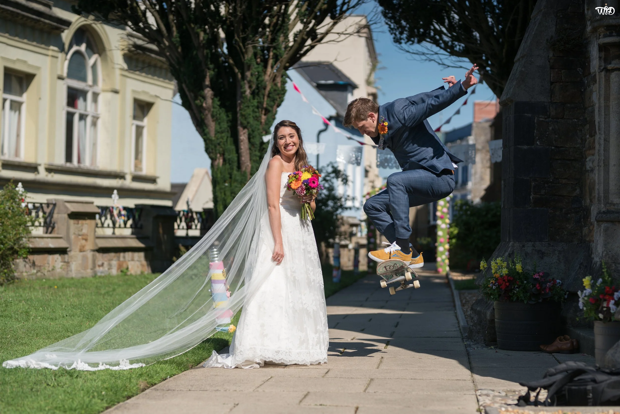 A bride in a white wedding dress with a long veil holding a bouquet of colorful flowers, smiling, while a groom in a blue suit and sneakers is skateboarding towards her, outdoors on a sunny day with historic buildings and decorative bunting in the ba