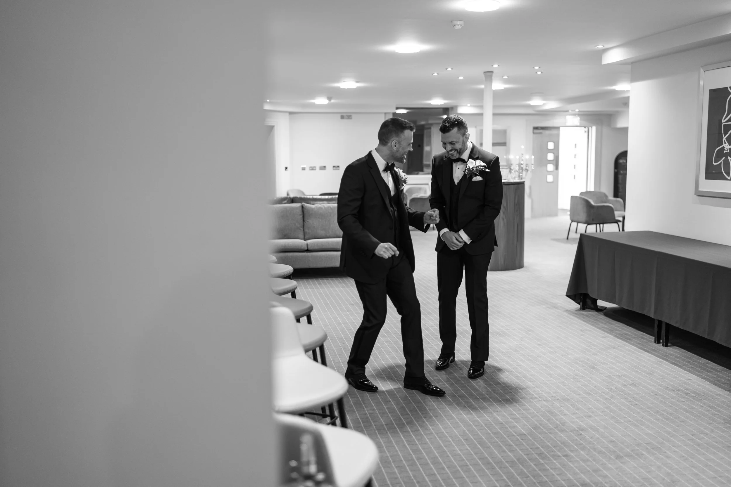 Two men in tuxedos sharing a moment of laughter in a hotel lobby.