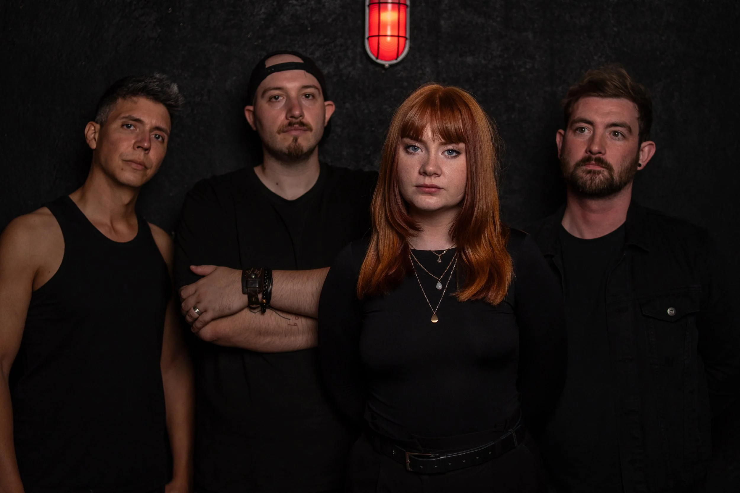 Four musicians standing against a black wall, with a red light fixture above them, posing for a band photo. Promotional Music Photography.