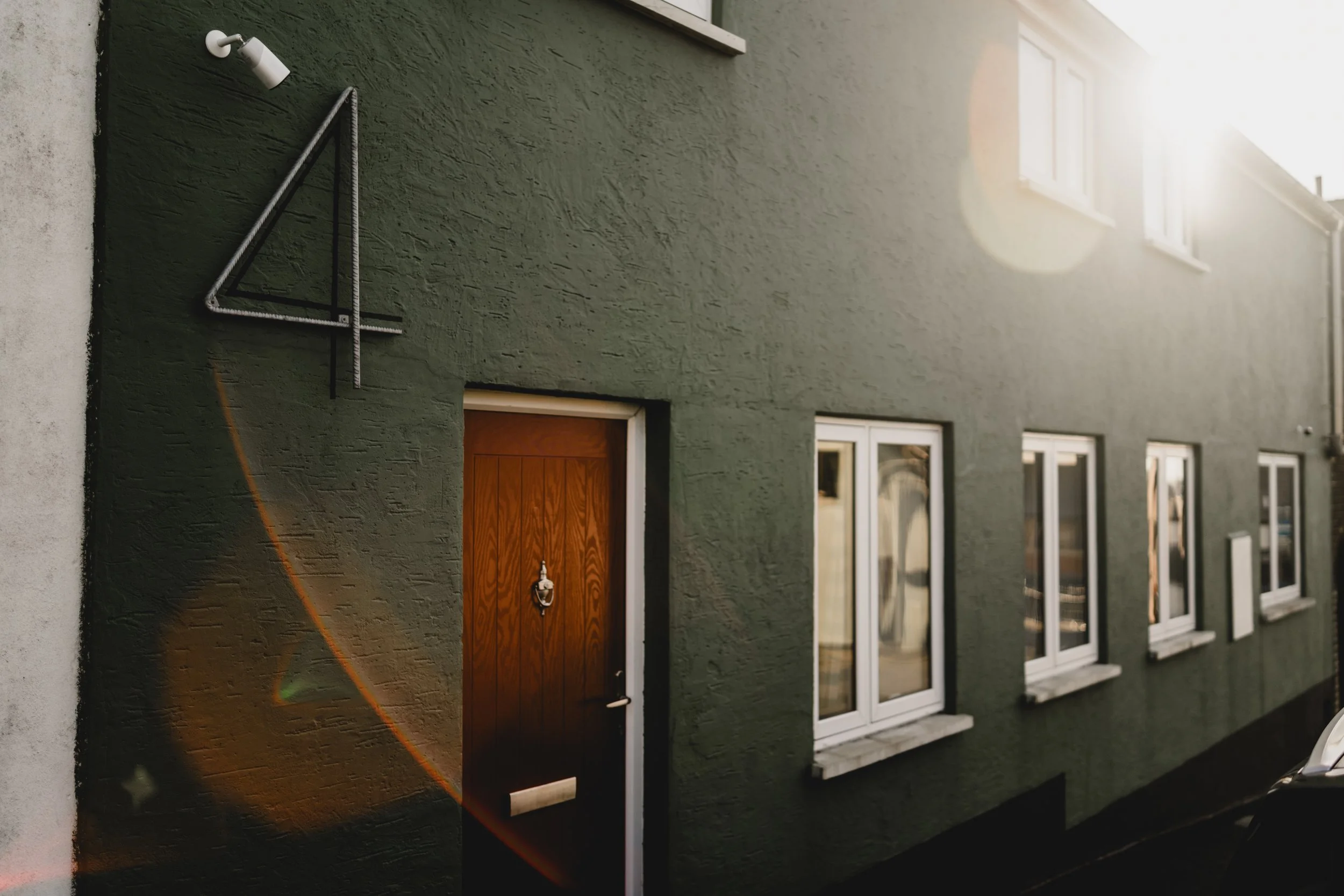 A green building with a wooden front door, several windows, and a triangular metal design element on the wall. The sunlight is shining from the top right corner, creating a lens flare.