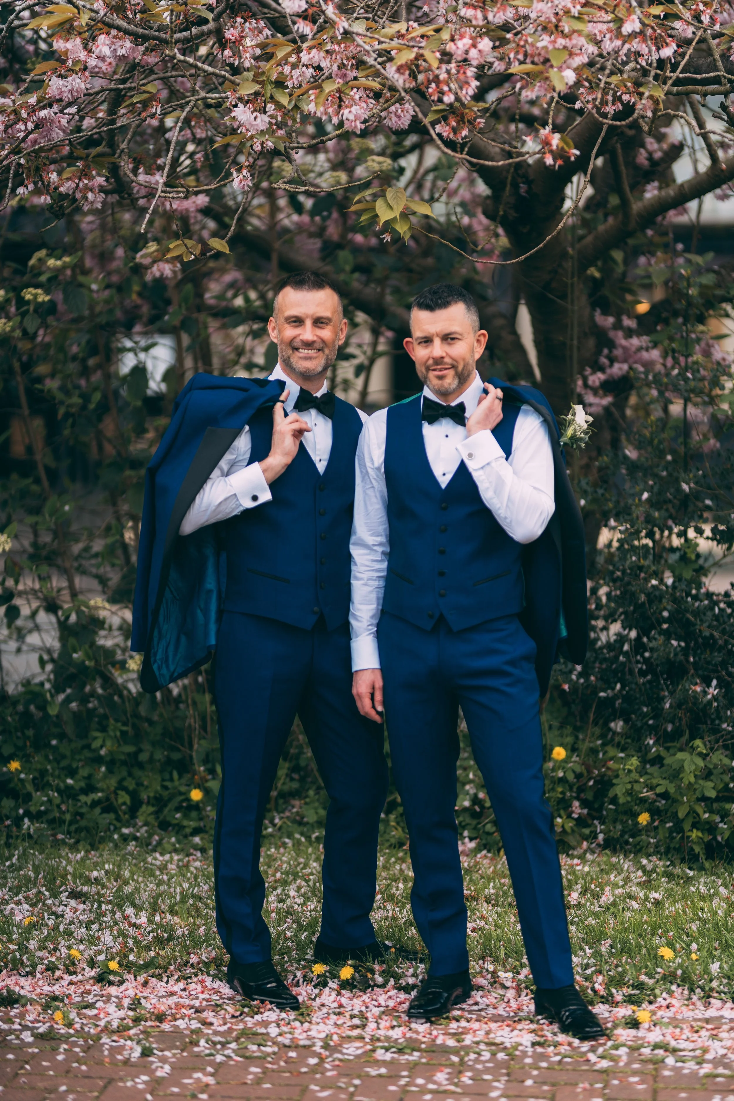 Two men in blue suits, white shirts, and black bow ties standing outdoors under a flowering pink cherry blossom tree, carrying matching jackets over their shoulders, smiling.