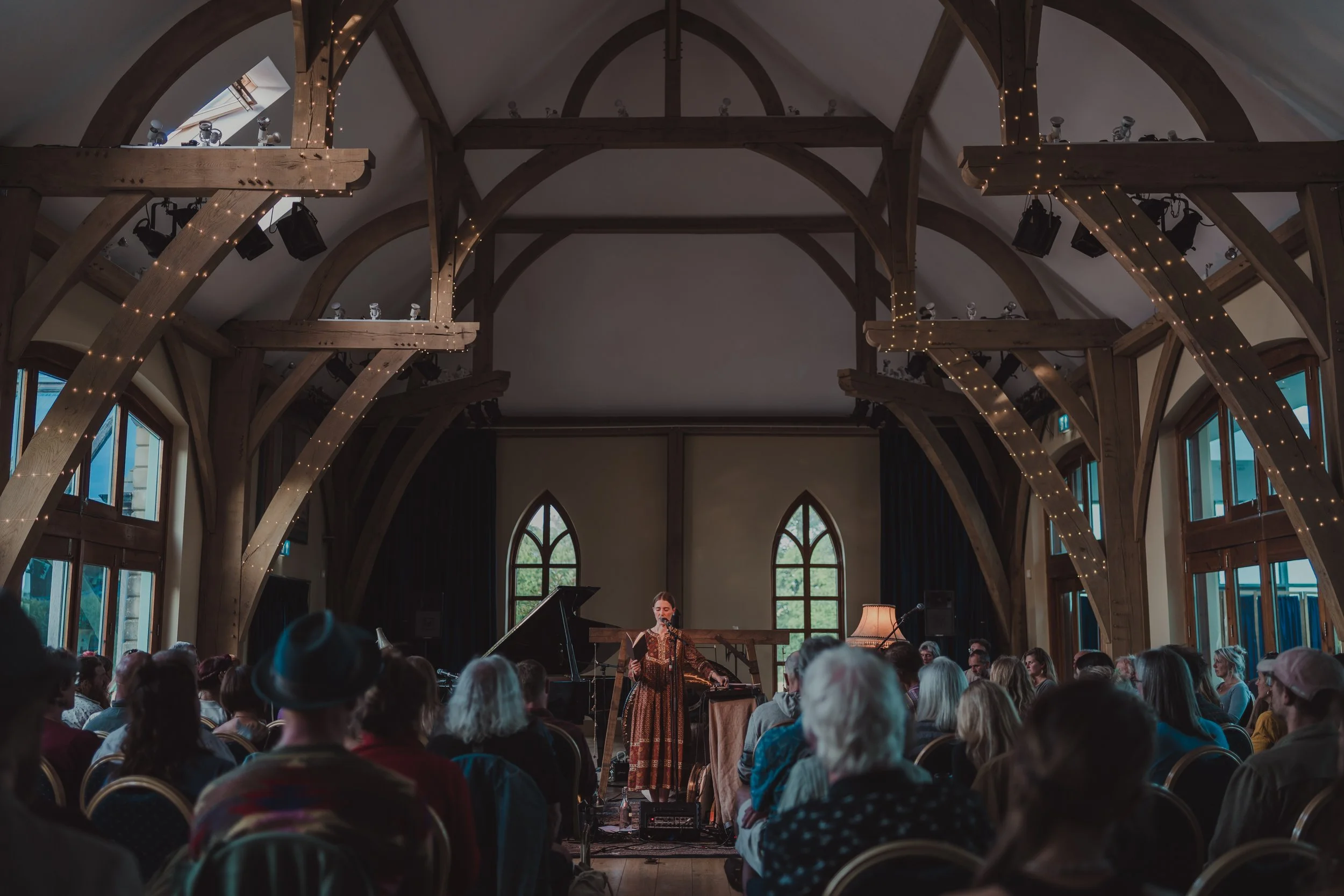 A woman performing on a stage with a microphone in front of an audience in a room with high, arched wooden beams and large windows.