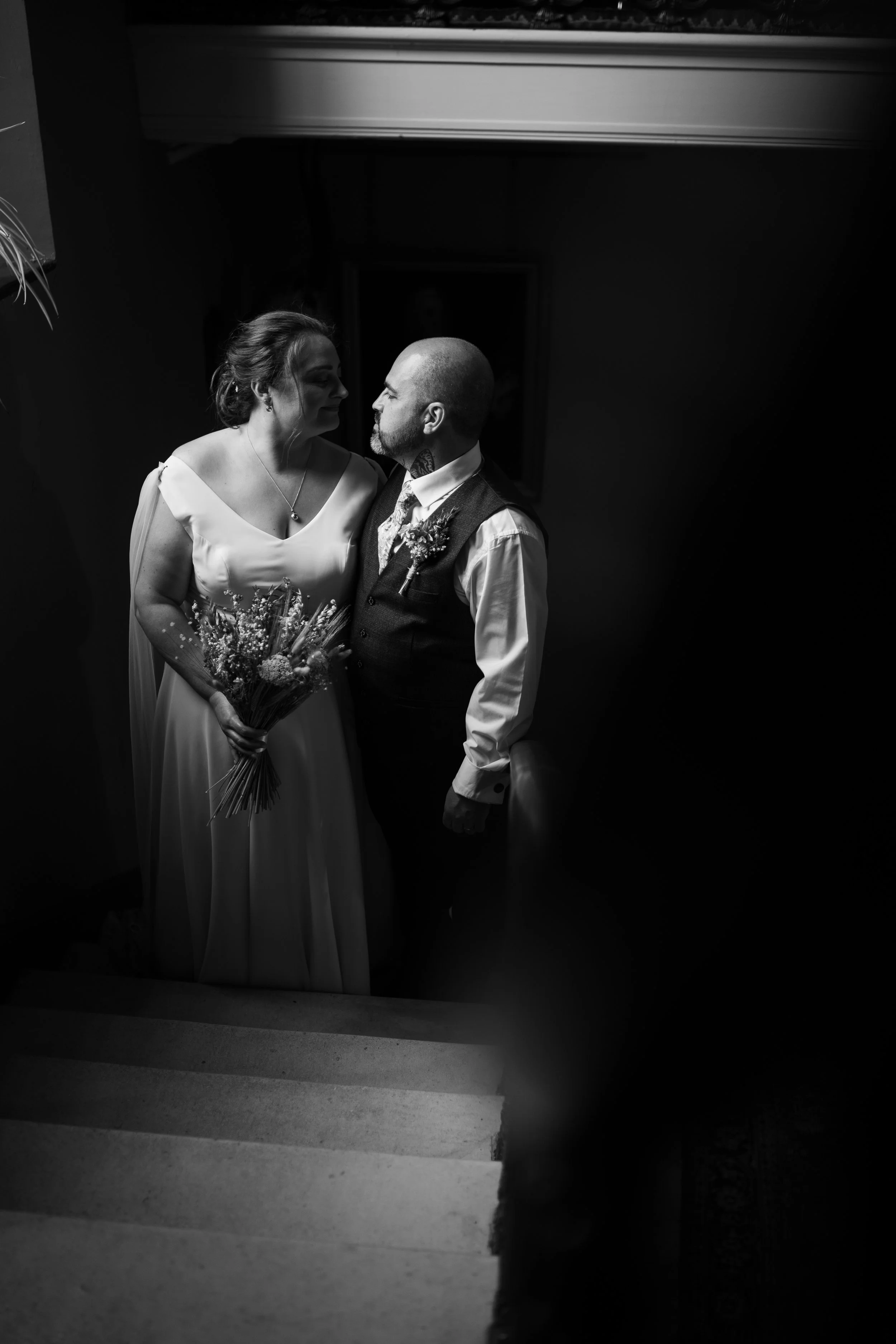 Black and white photo of a bride holding a bouquet, and a groom standing close to her, looking into her eyes.