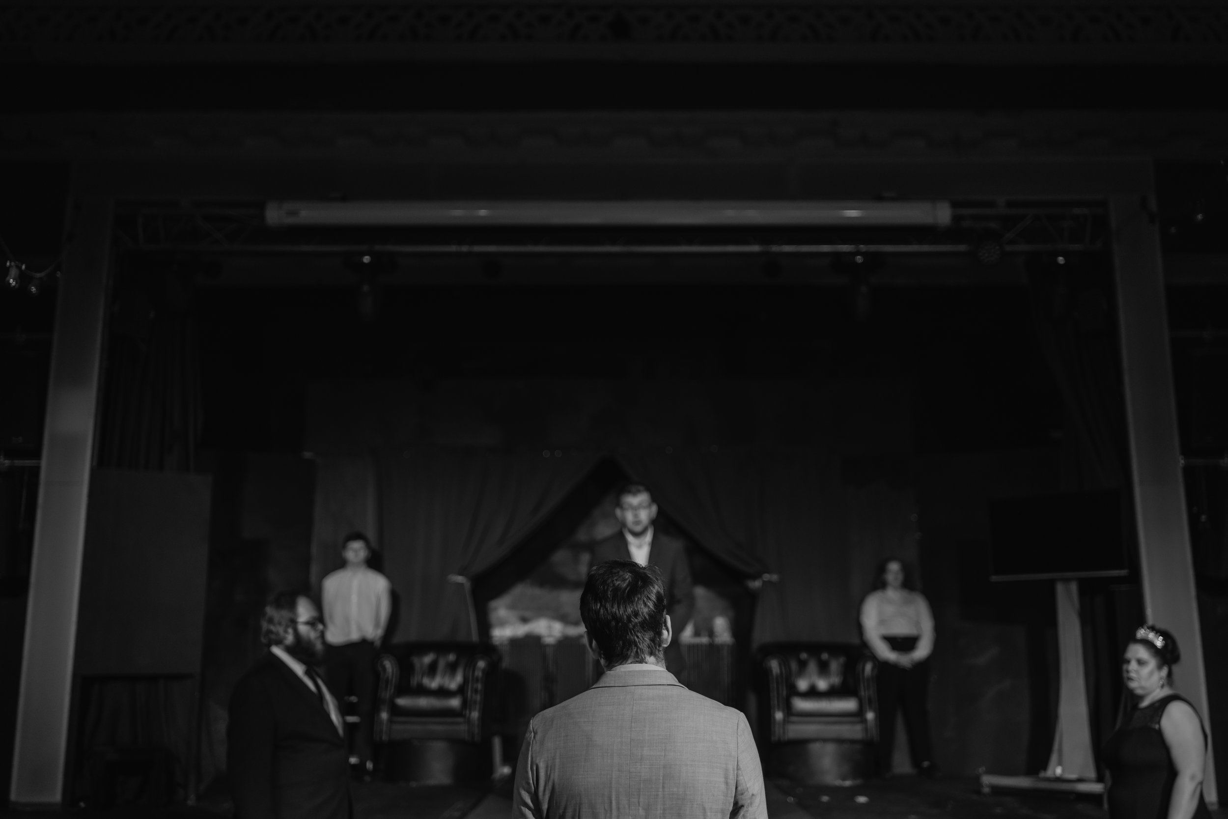 A black and white image of a theater stage with five people. Four are standing at different points and one person is seated behind a curtain with a landscape backdrop. The stage has two chairs, and a curtain in the background.