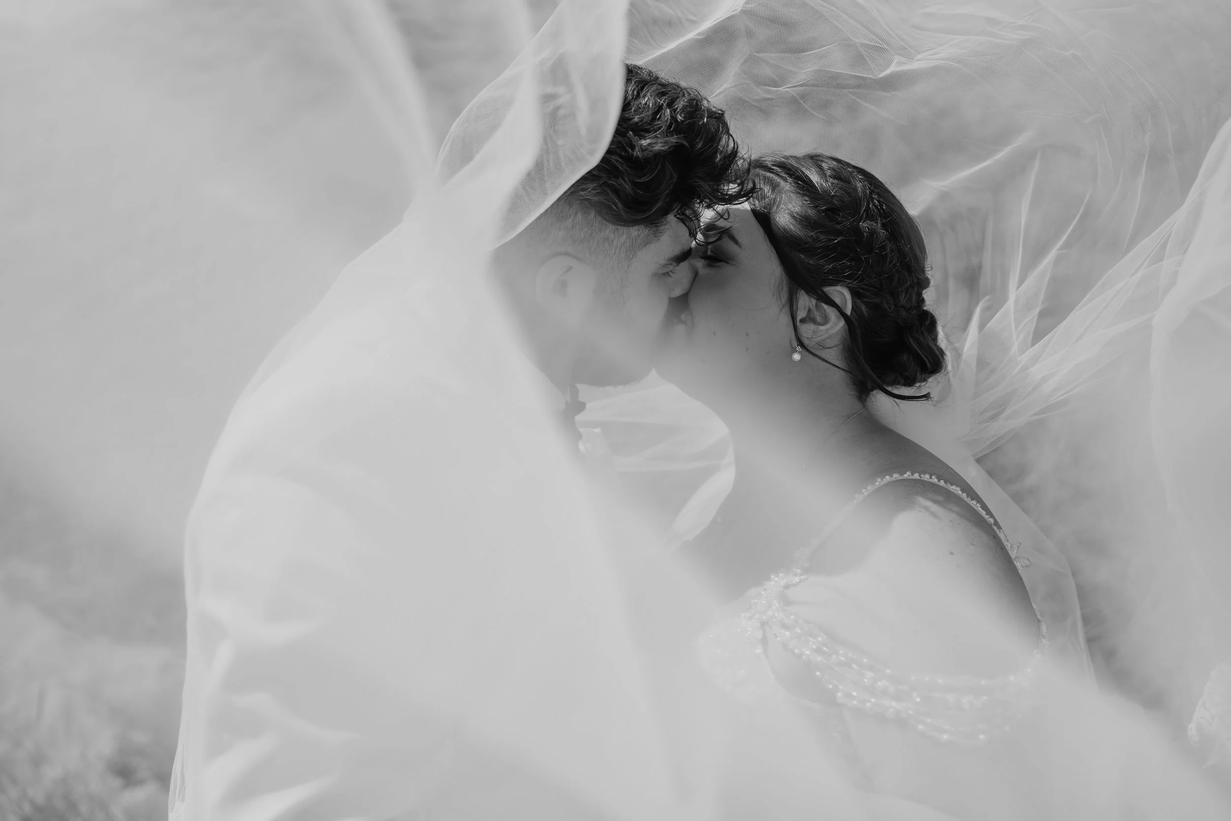 A black-and-white photo of a couple kissing, partly covered by a sheer fabric, with one person wearing a wedding dress and the other in formal attire.