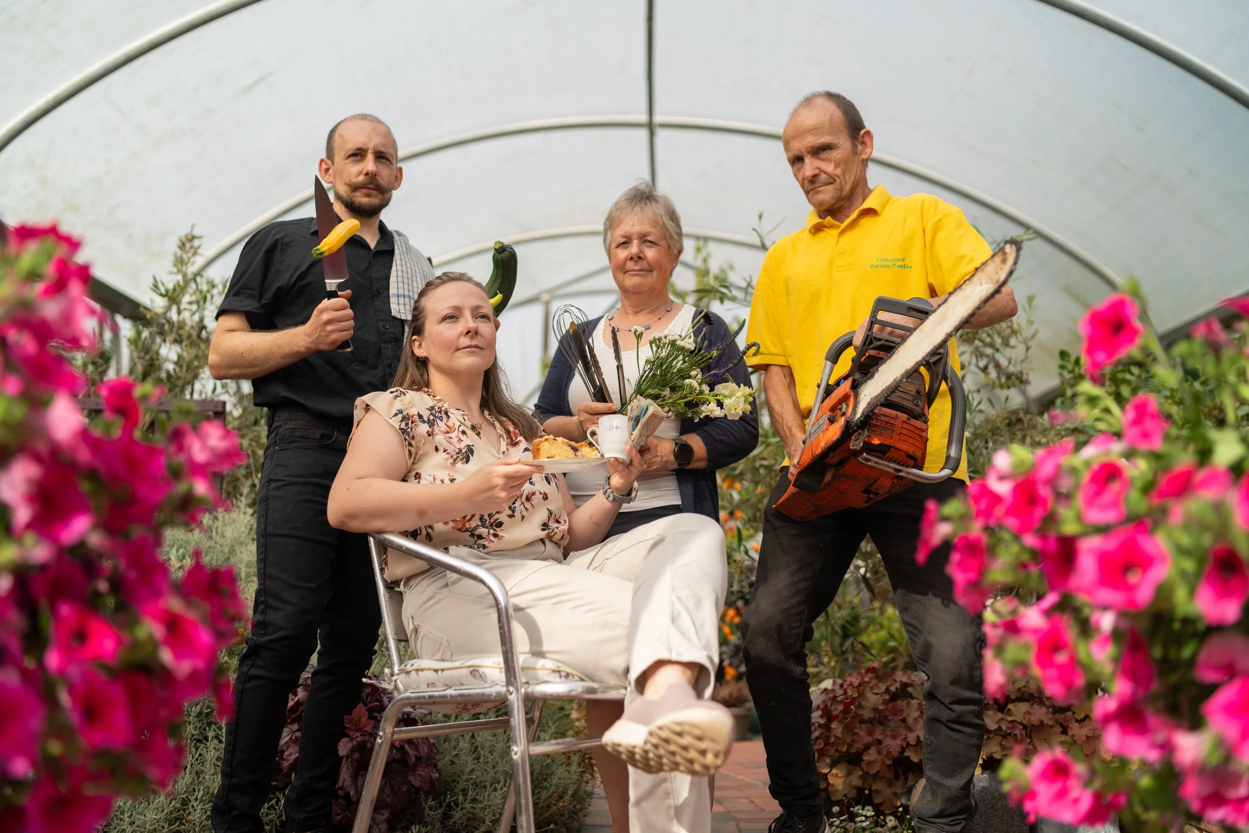 A group of five people in a greenhouse, with one woman seated in a chair, holding a plate of food, surrounded by plants and flowers.