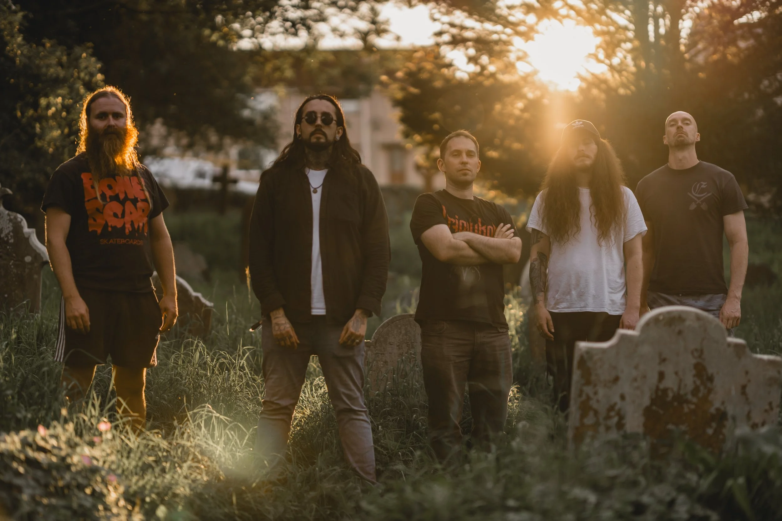 A group of five men standing in a grassy area with tombstones, during sunset, with trees in the background. Promotional Music Photography.