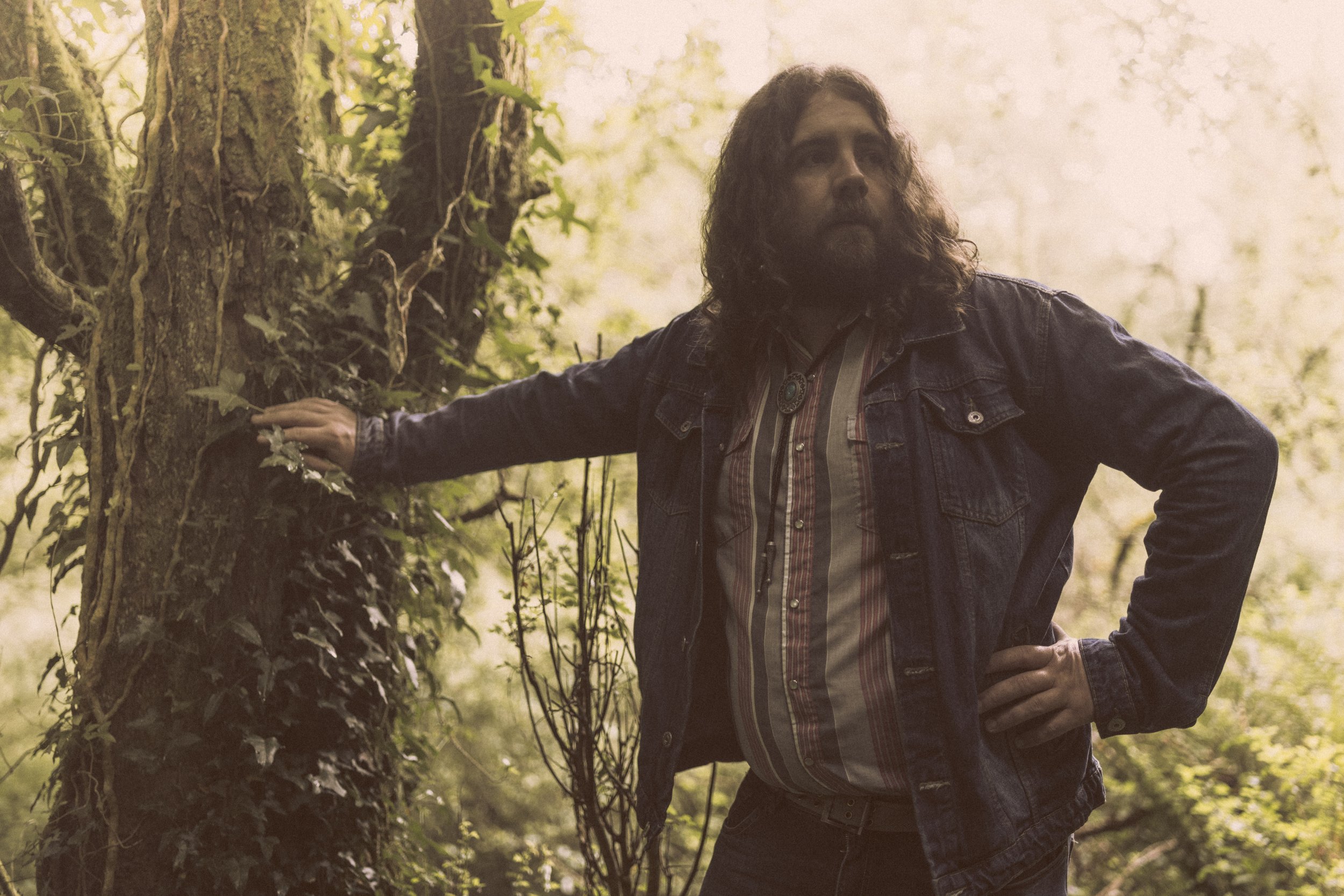 A man with long curly hair and a beard standing in a forest, leaning against a tree with one hand on his hip and the other resting on the tree. Promotional Music Photography.