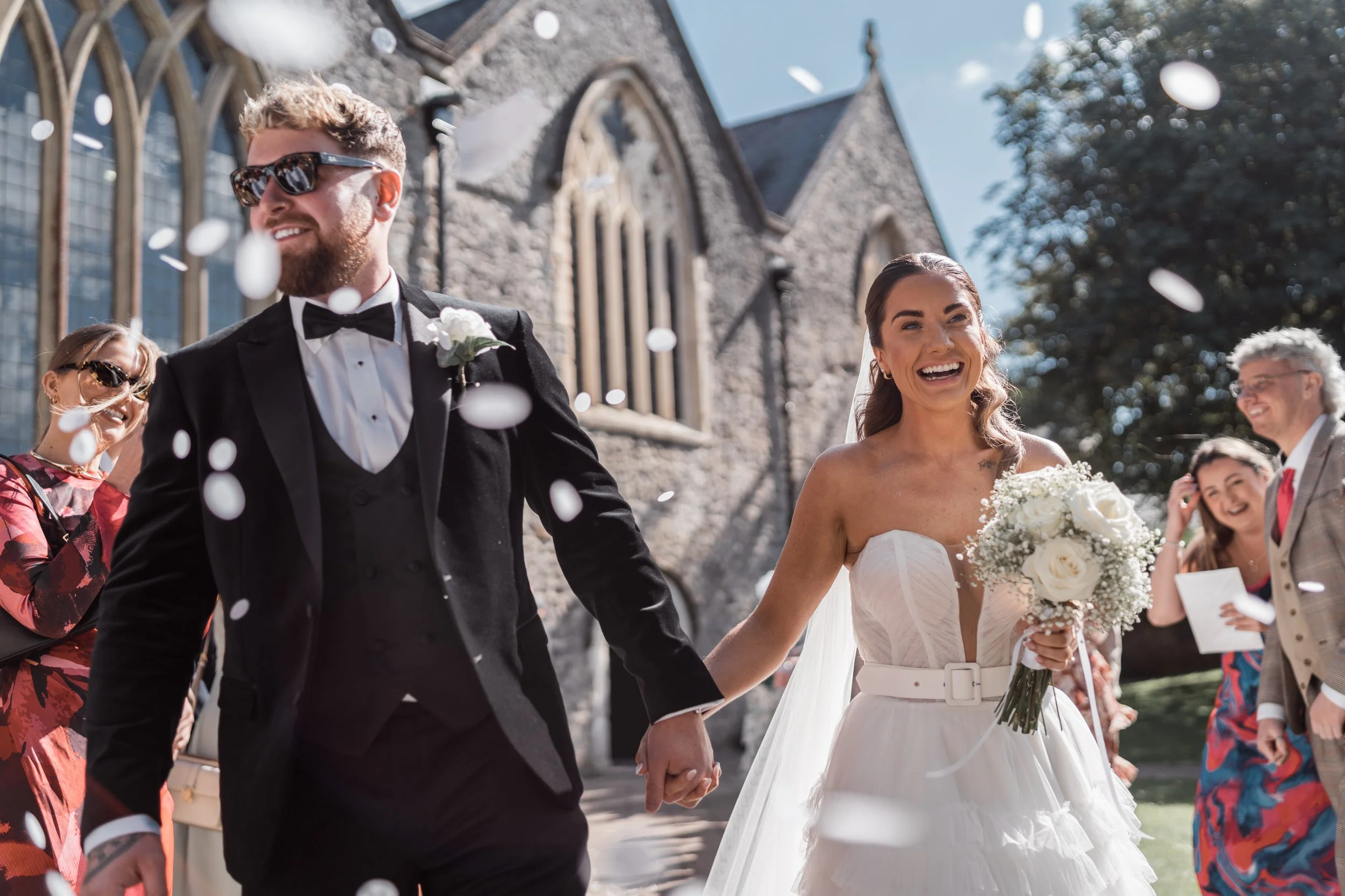 A bride and groom walking hand-in-hand outside a church, celebrating their wedding with guests and falling confetti.