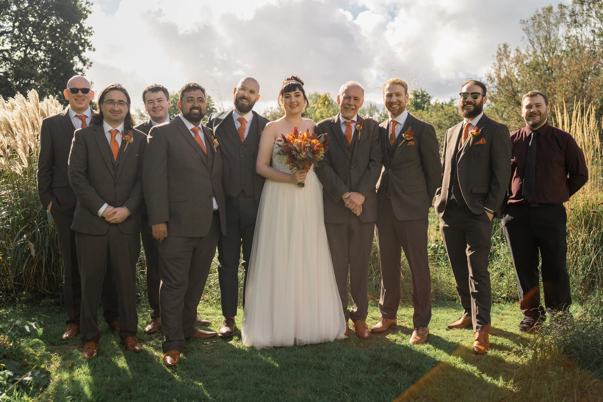 A group of people in suits and dresses standing outdoors, smiling, during a wedding. The bride in a white dress is holding a bouquet of flowers, and there are men in brown suits with orange accents. The background has trees and tall grass.