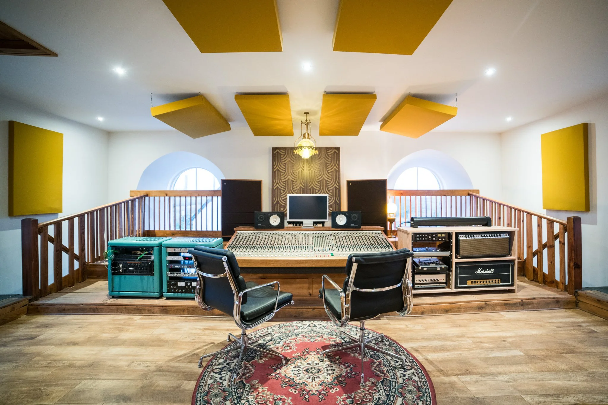 Recording studio with a large mixing console, two chairs, audio equipment, and soundproofing panels on the ceiling.