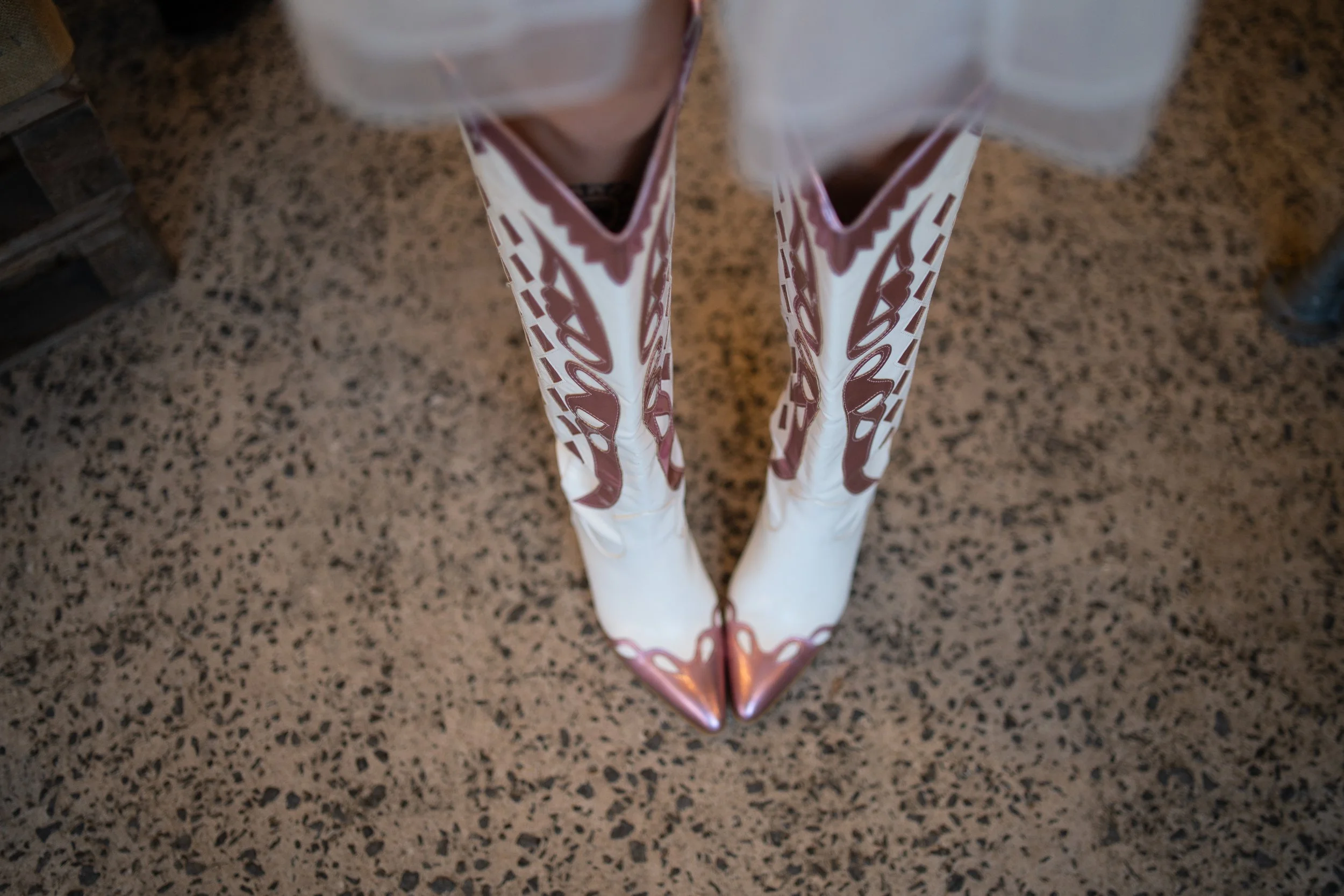 Pair of white and pink cowboy boots with pointed toes, seen from above on a speckled stone floor.