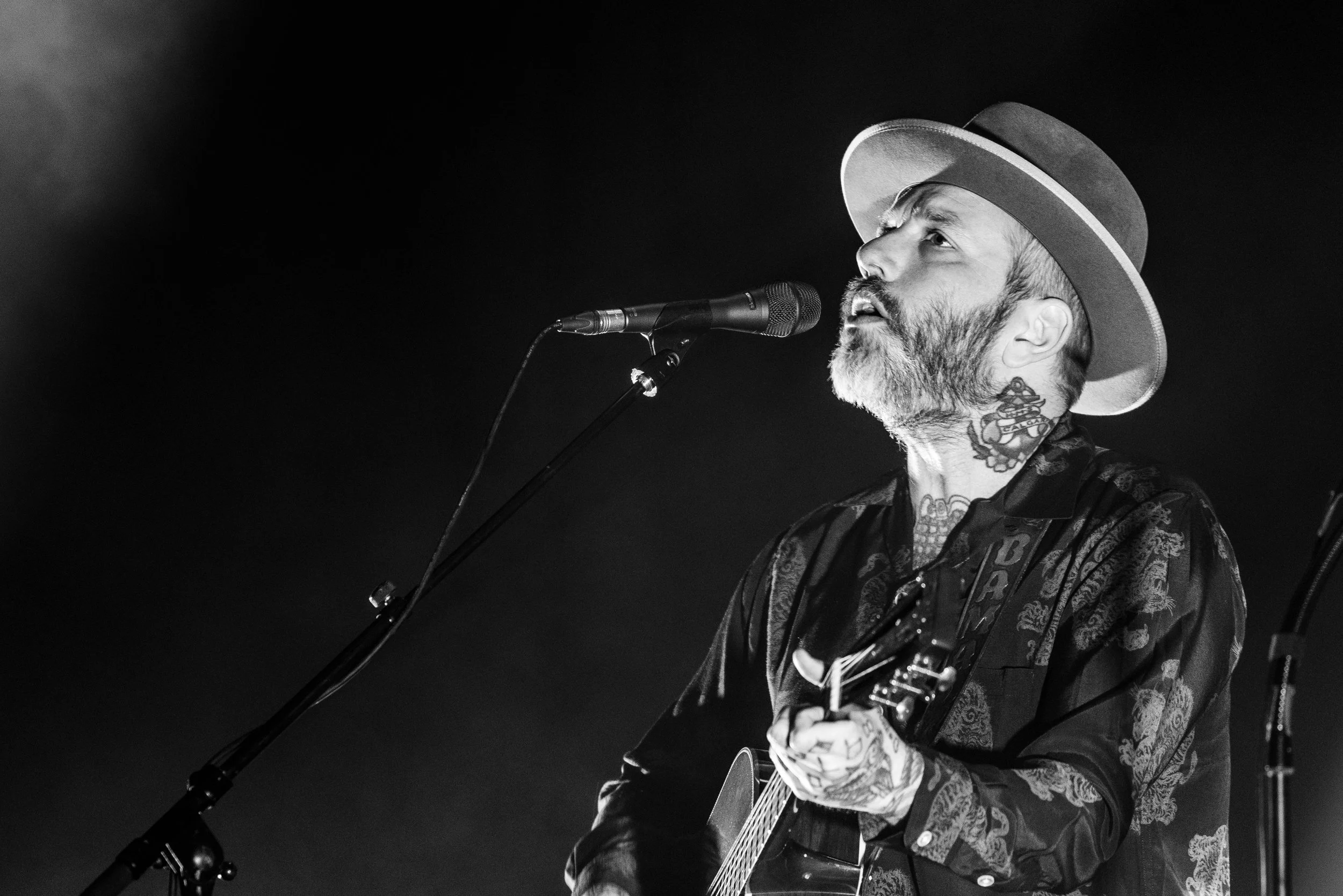 A black and white photo of a man singing into a microphone while playing an acoustic guitar, wearing a hat and a patterned shirt. City & Colour musician Dallas Green.