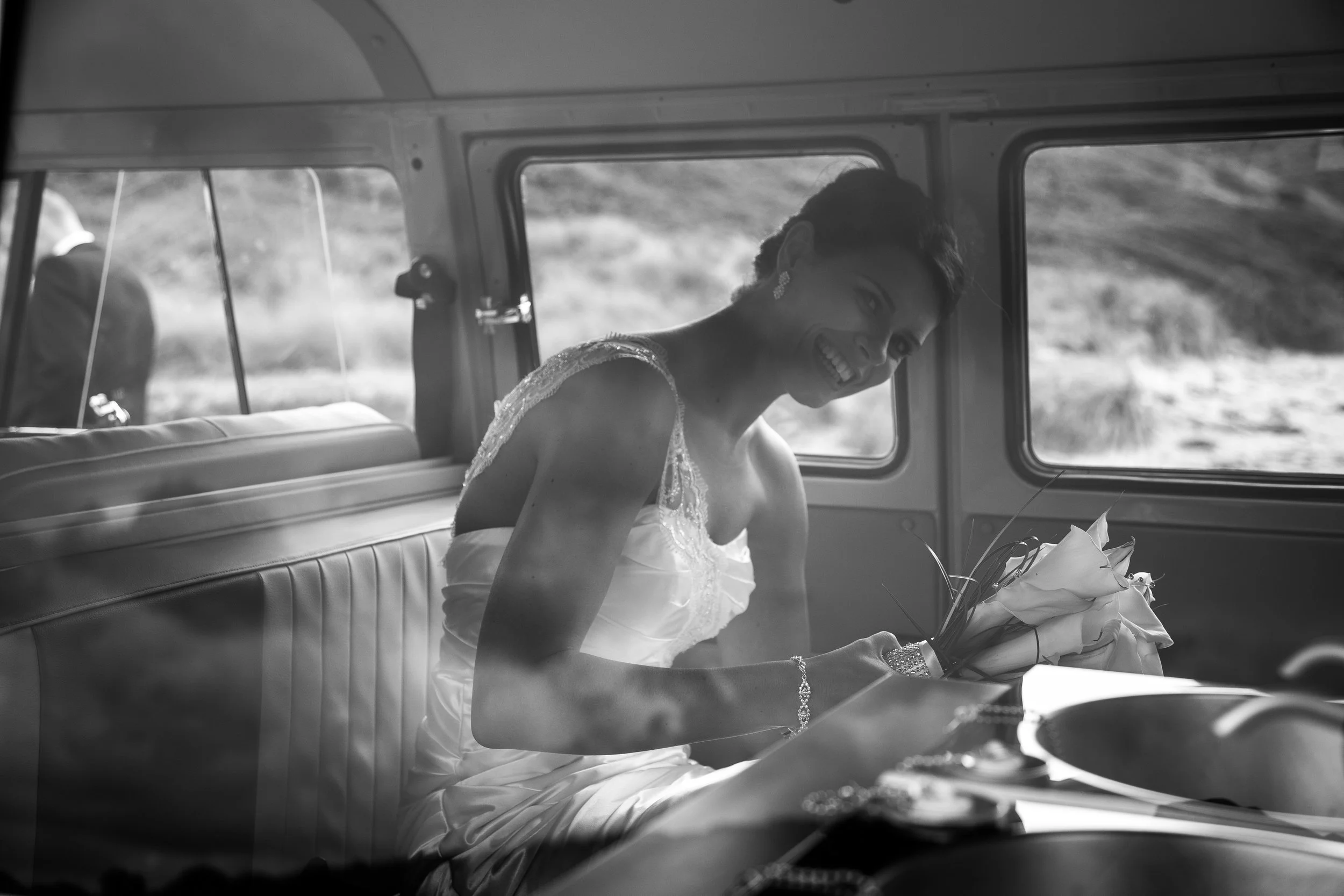 A woman in a wedding dress sitting inside a vehicle, holding a bouquet of flowers, smiling and looking at the camera.
