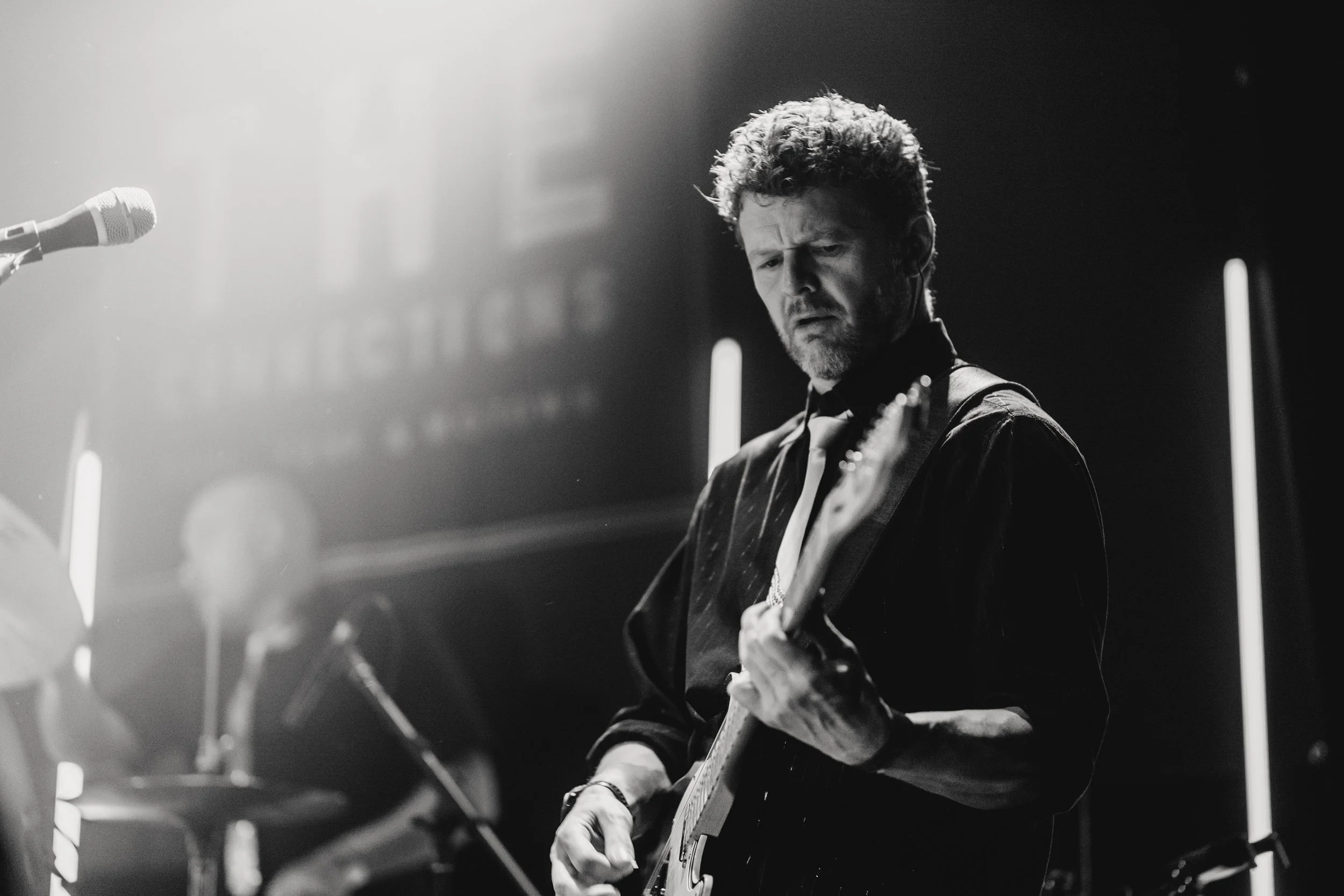 A black and white photograph of a man playing an electric guitar on stage, with a focused expression on his face, and a blurred musician playing drums in the background. A microphone is visible on the left side. Live Event Photography.