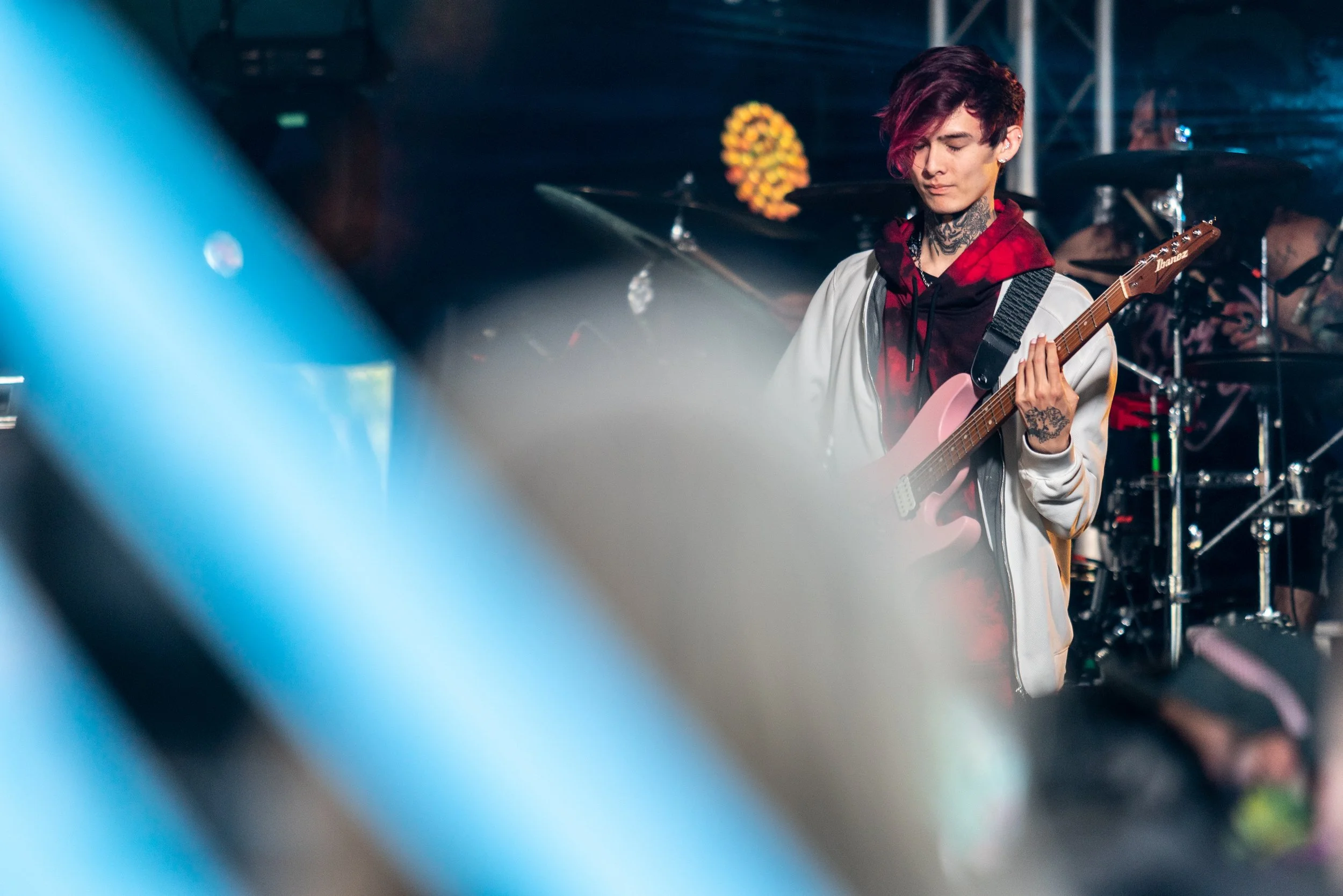 A young man with short, dark red hair playing an electric guitar on stage during a concert, with drum set and other band members in the background. Polyphia Band.