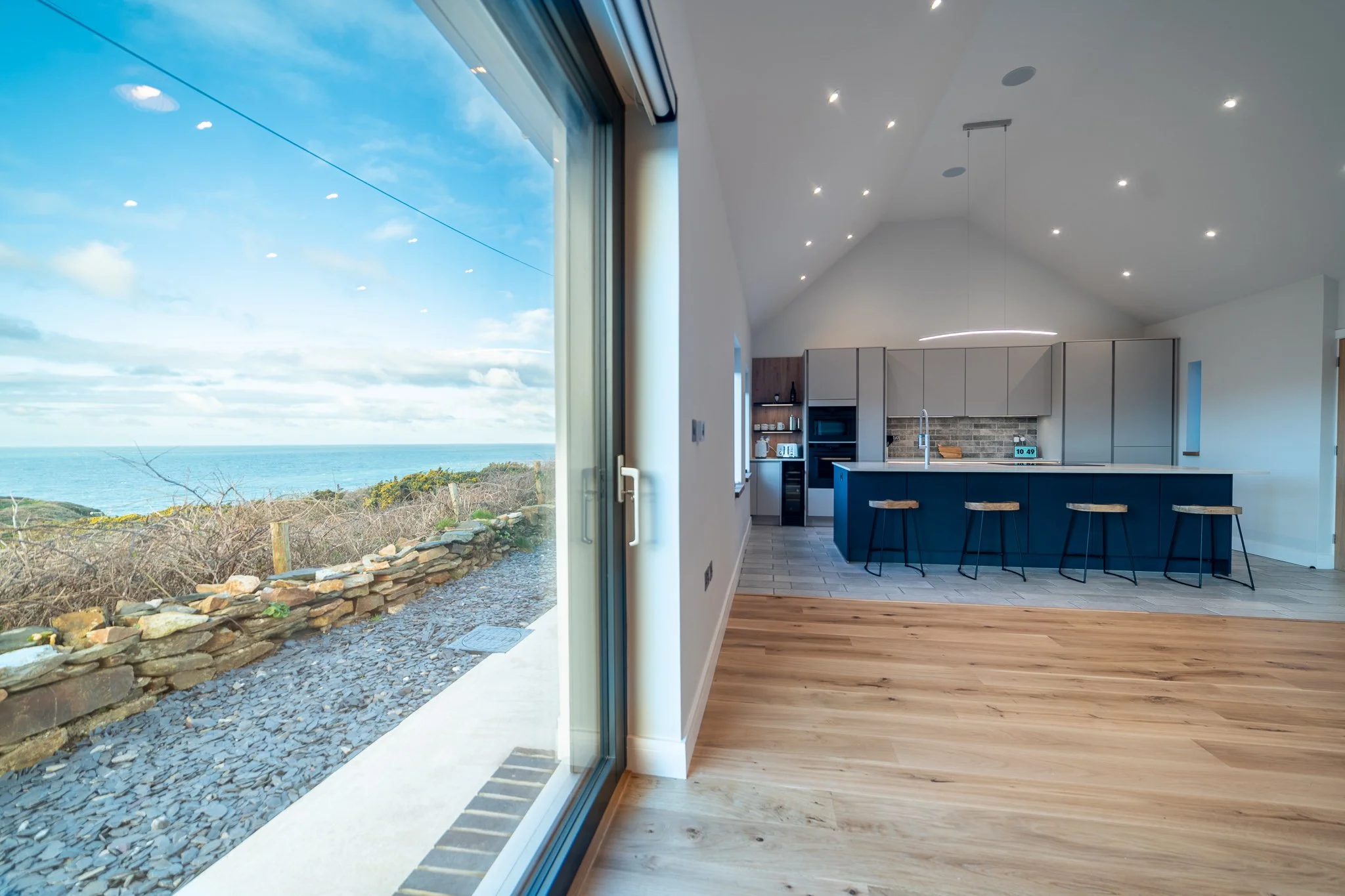 Modern open-concept kitchen with a view of the ocean through a large glass sliding door, featuring a blue kitchen island with bar stools, wood flooring, and a sky with clouds outside.