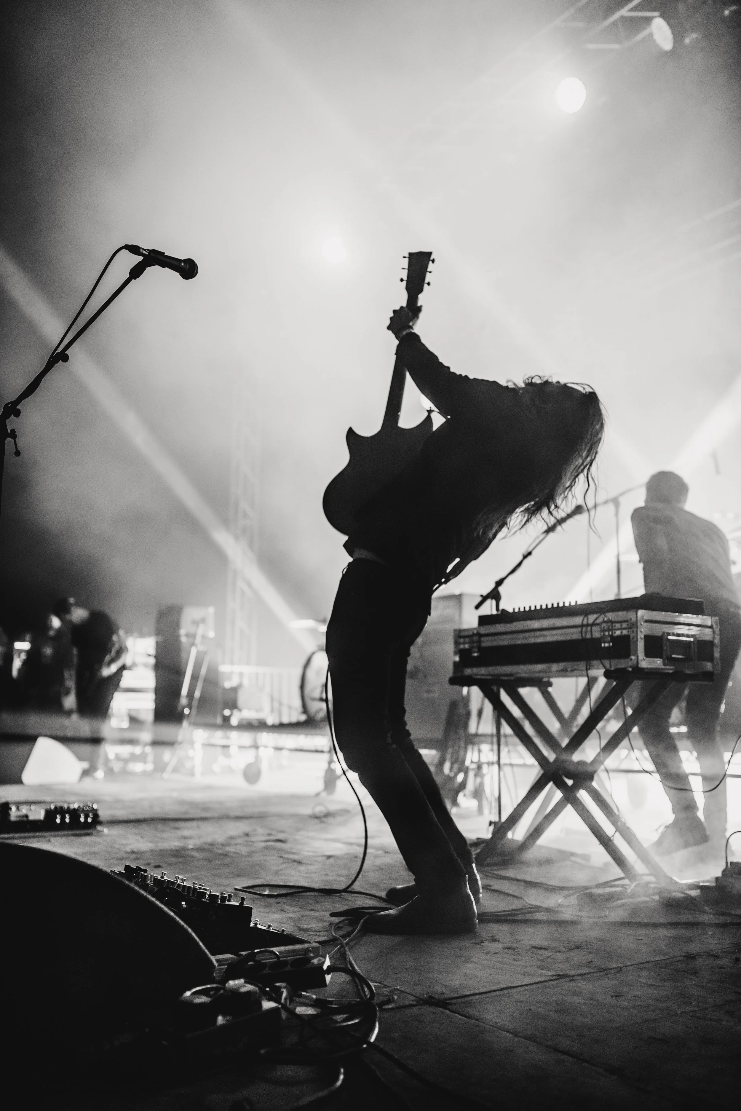 Silhouette of a guitarist performing on stage with a keyboard next to them, backlit by stage lights, in a concert setting.
