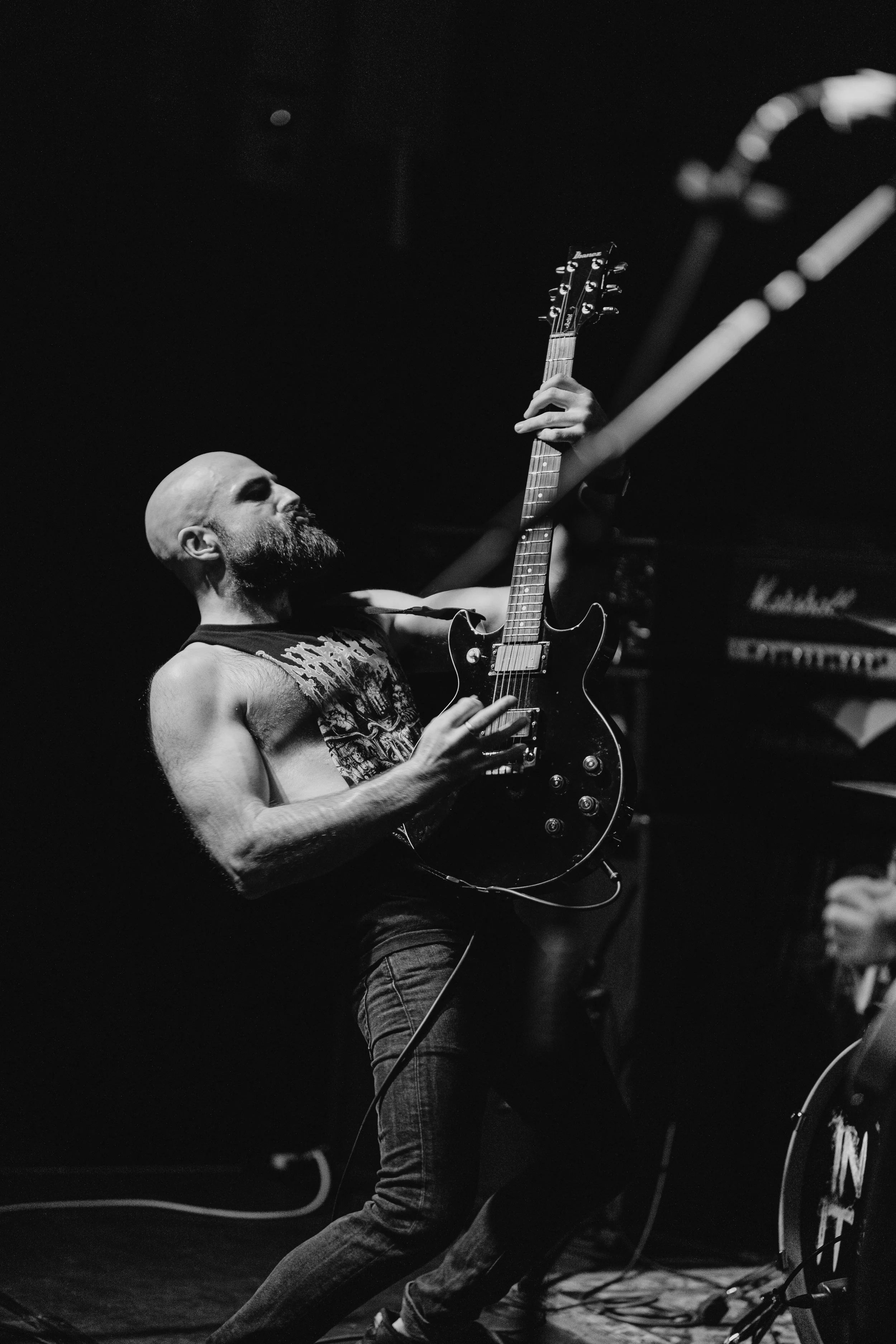 A bald man with a beard, wearing a sleeveless graphic shirt, is playing an electric guitar on stage, with a Marshall amplifier visible in the background. Live Event Photography.