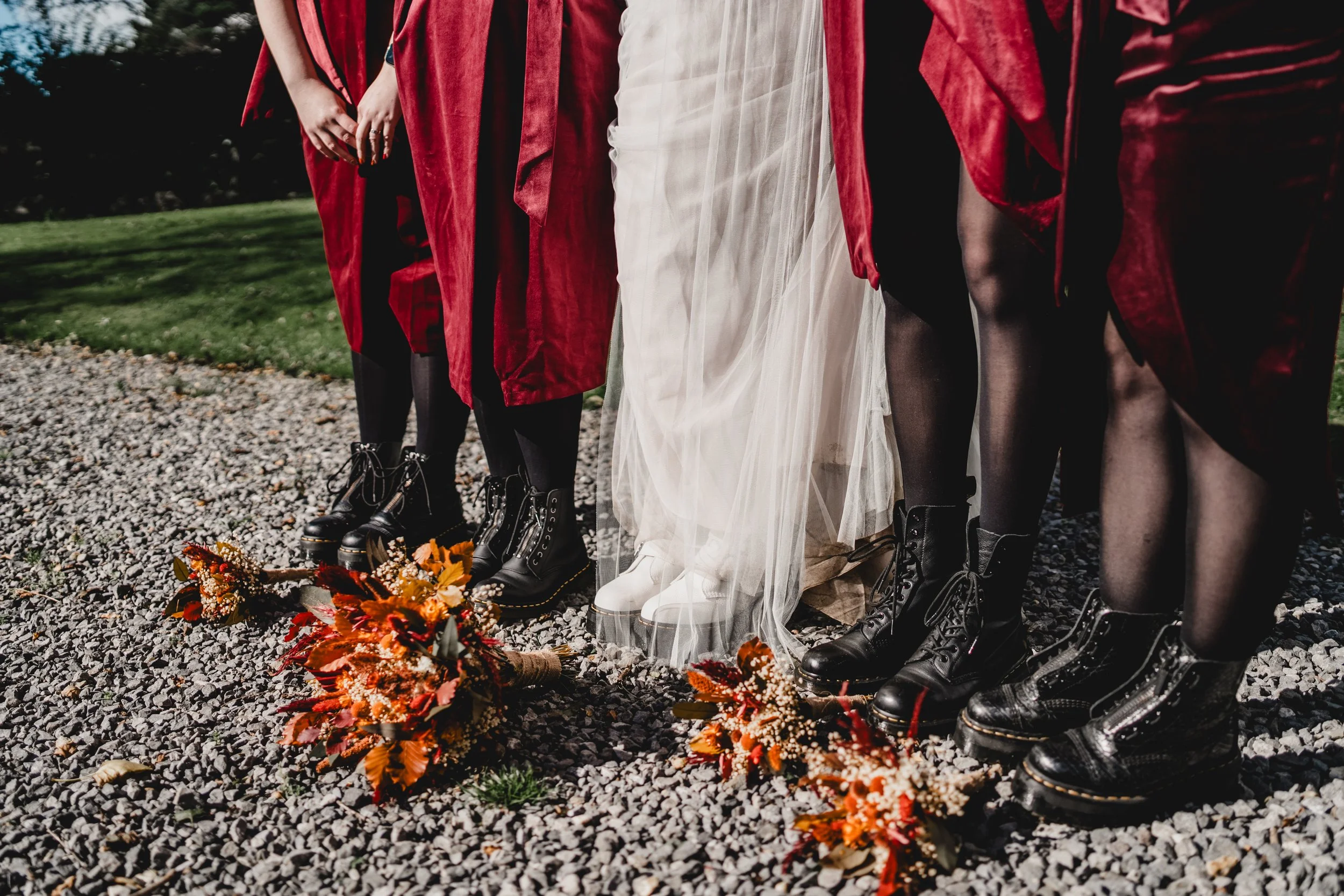 People standing outdoors in black combat boots, with a bouquet of orange and red flowers on the gravel ground, during a wedding or similar celebration.