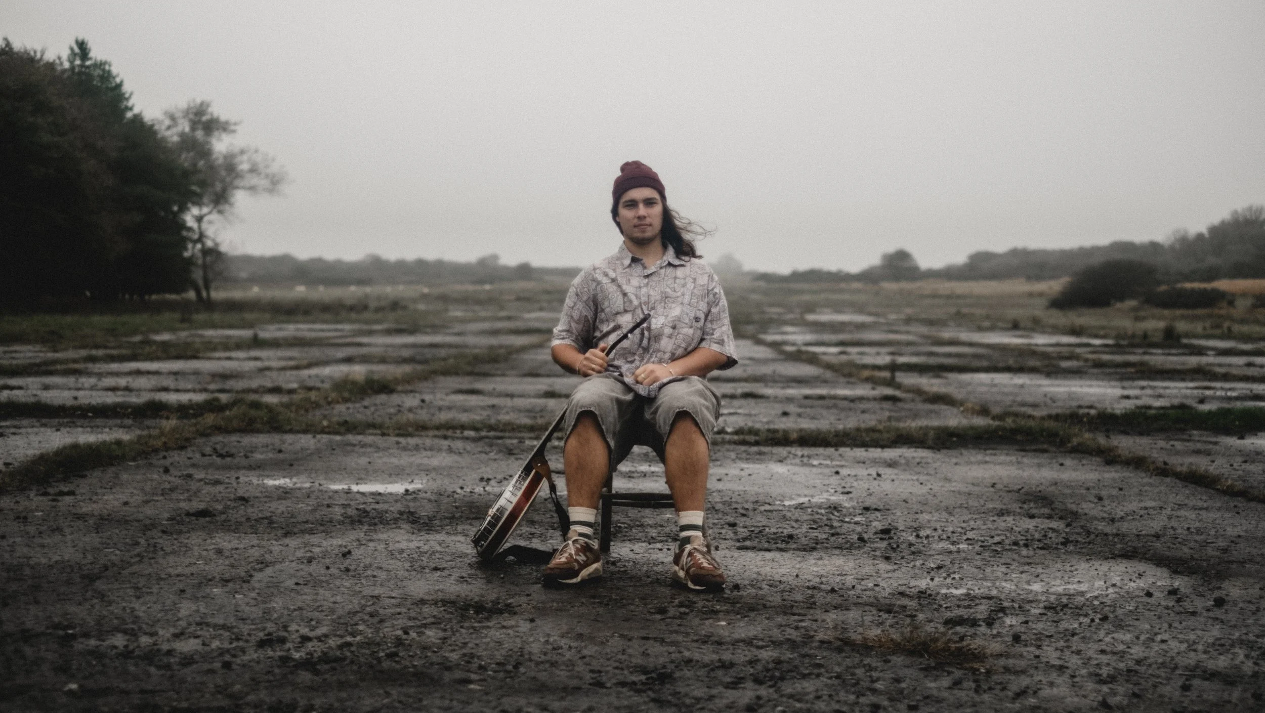 A young man sitting on a small chair on an empty, wet, and rugged concrete surface outdoors, holding a guitar and wearing a knitted cap, a patterned shirt, shorts, and sneakers, with trees and an overcast sky in the background. Promotional Music Phot