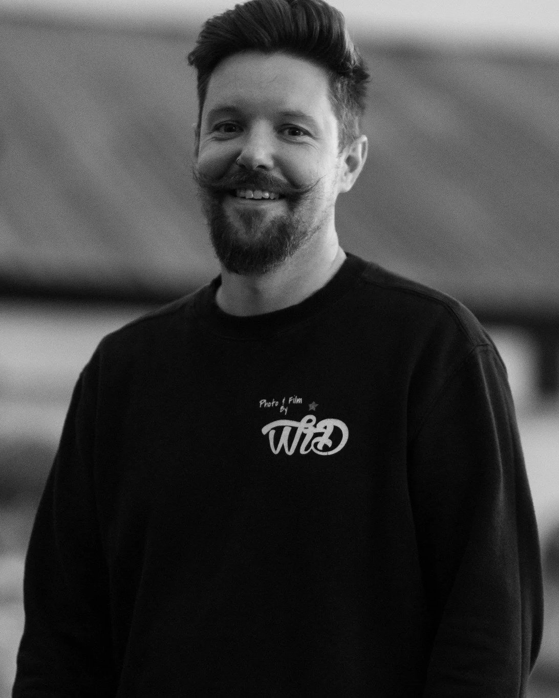 A smiling man with a beard and styled hair wearing a black sweatshirt with white WetheDee logo on the chest.