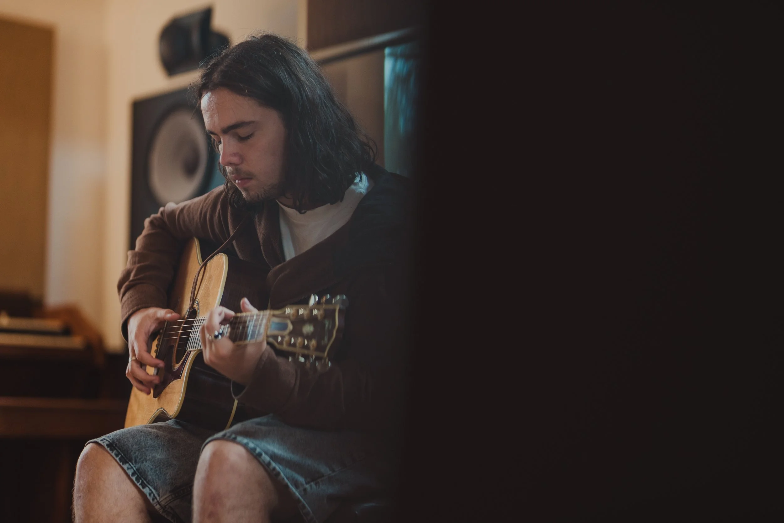 A young man with long dark hair, wearing a brown hoodie and denim shorts, sitting and playing an acoustic guitar in a room with musical equipment.
