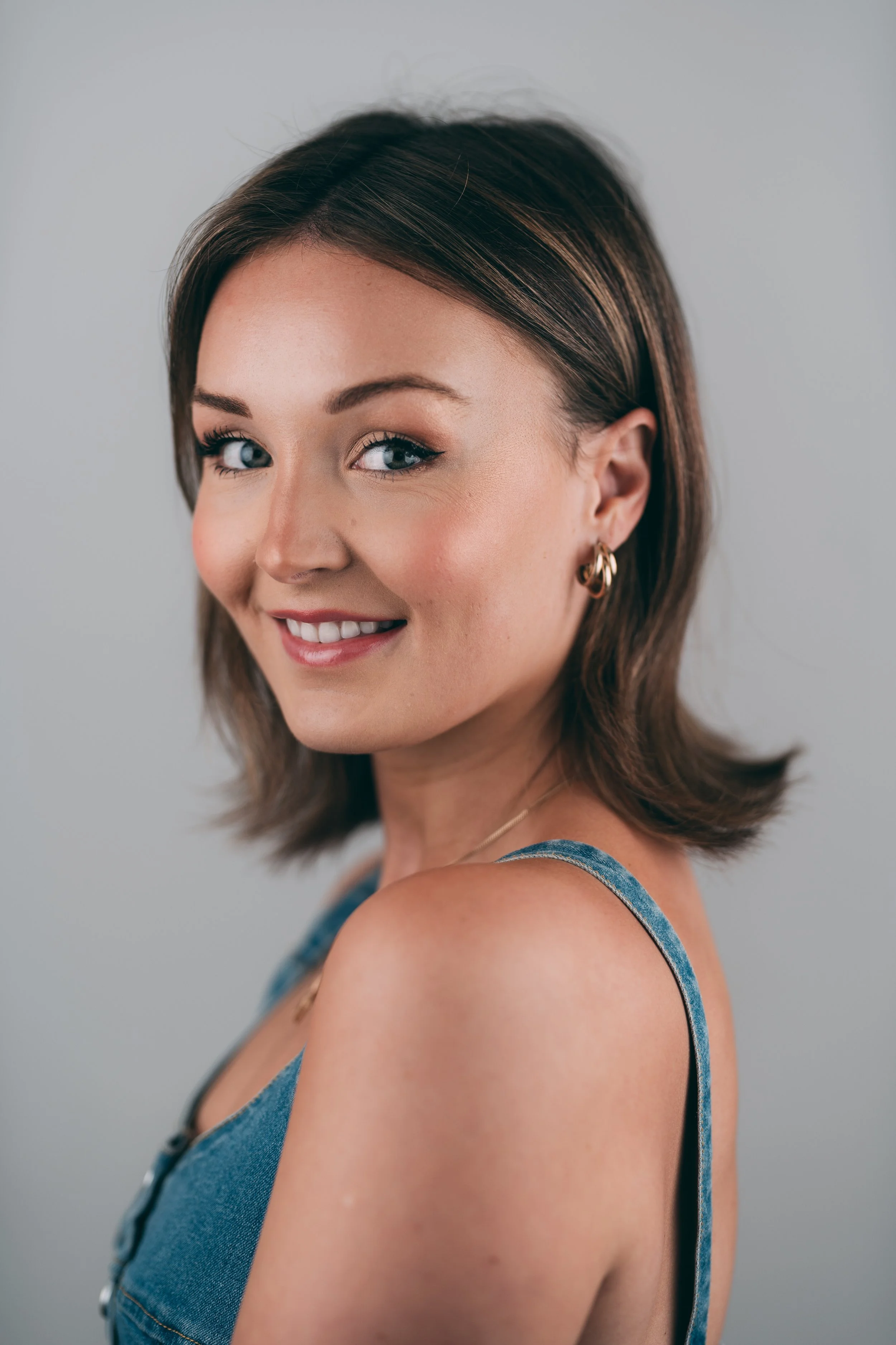 A young woman with medium-length brown hair, blue eyes, and earrings, smiling at the camera. She is wearing a blue denim top and standing against a neutral background.