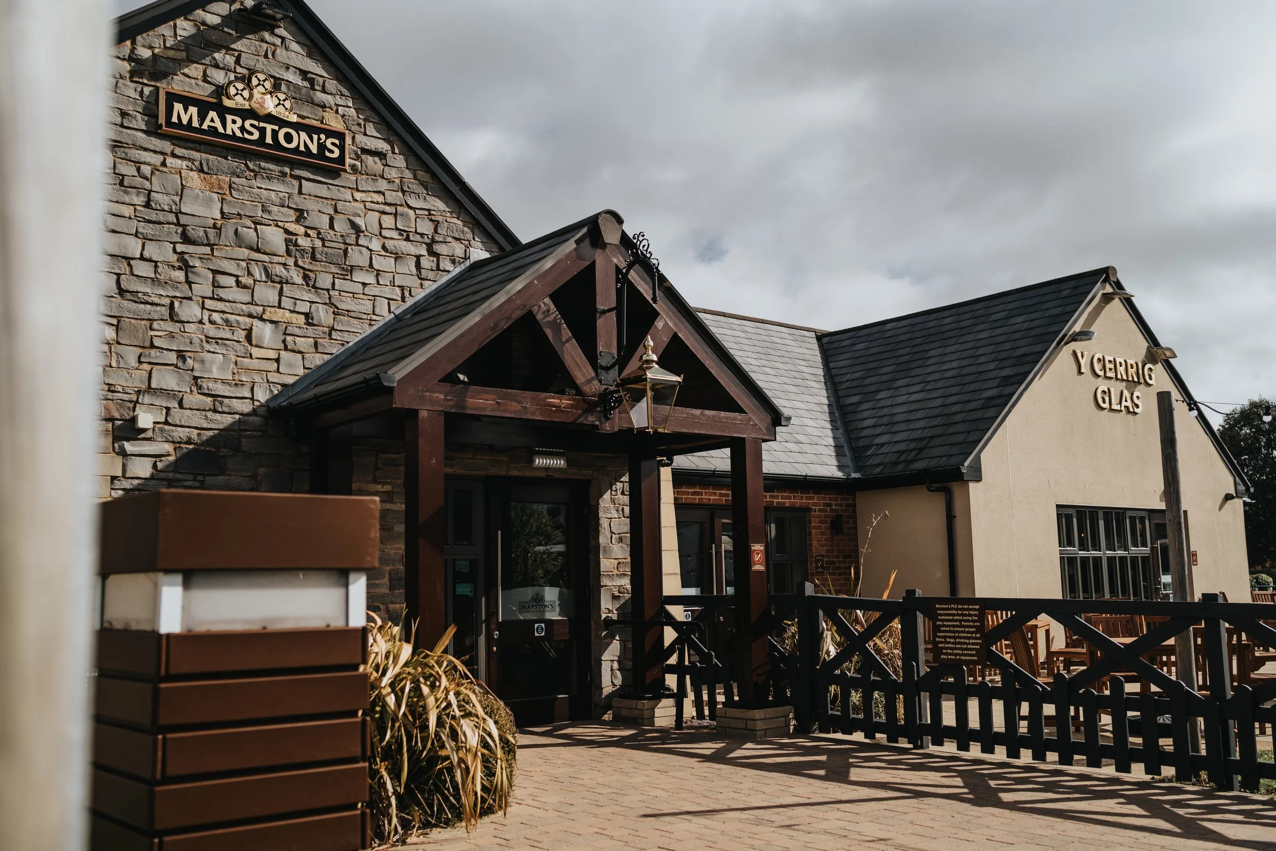Exterior of a building with stone and stucco walls, signage for 'Marston's' and 'Y Cerrig Glas'. The entrance features a small porch with a wooden overhang, lantern, and ramp. Outdoor seating area is visible to the right, under a cloudy sky.