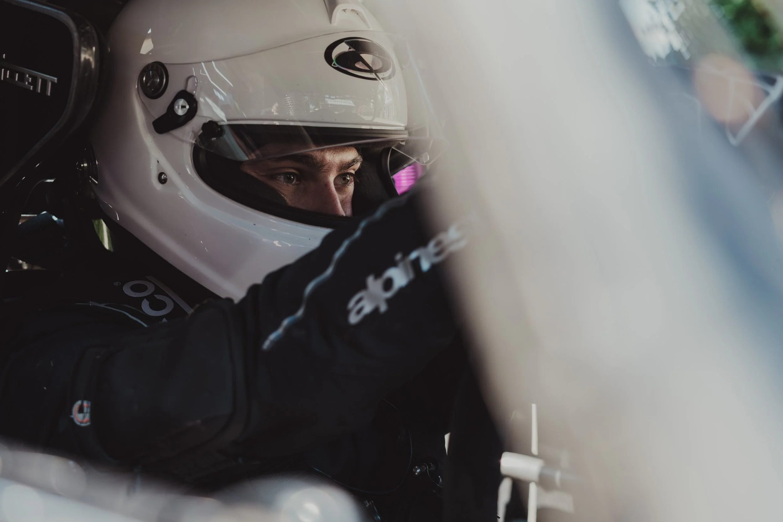 Close-up of a race car driver wearing a white helmet and black racing suit, focusing intently while sitting in the cockpit. Max Fewtrell of Quadrant.