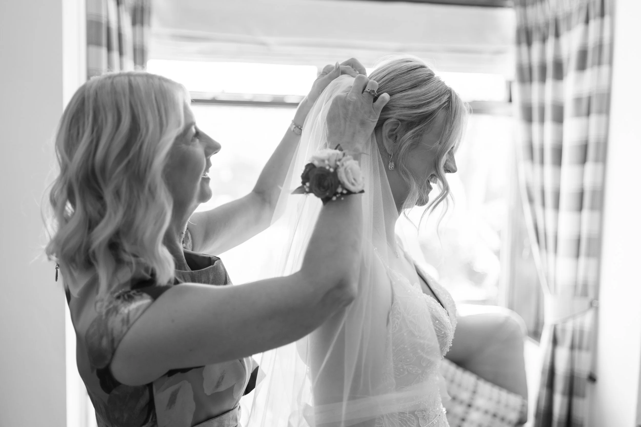 A woman helping a bride adjust her veil inside a room with curtains and sunlight.