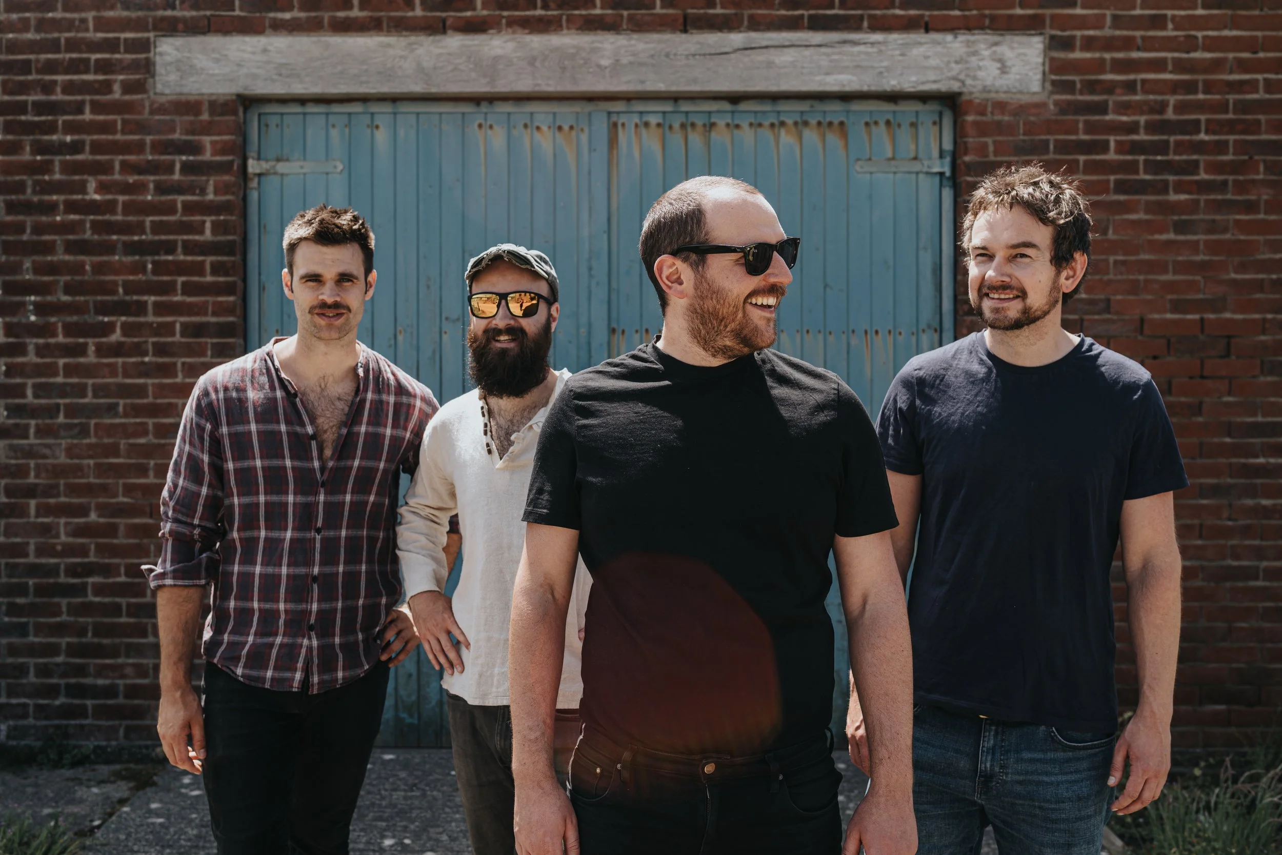 Five men standing in front of a brick wall and a blue garage door. They are smiling and looking off to the side, wearing casual clothing. Promotional Music Photography.