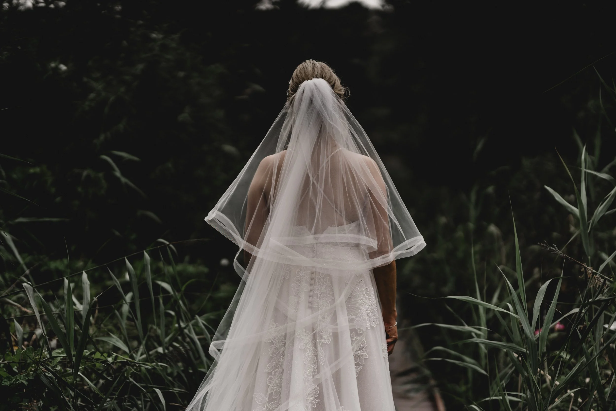 A woman in a wedding dress with a veil, viewed from the back, walking through a dark, lush outdoor setting. Alt Wedding. Alternative wedding photography.