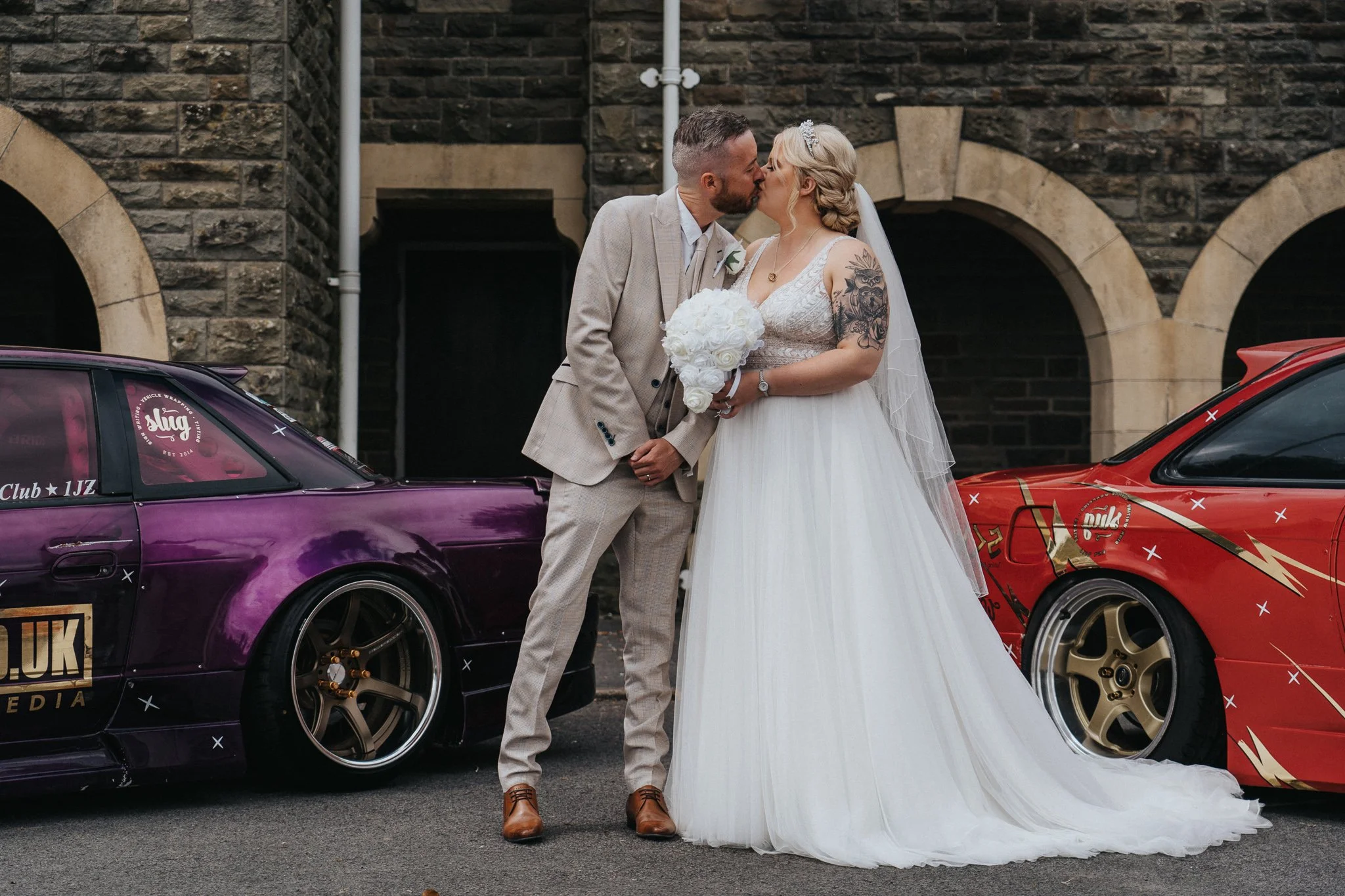 A newlywed couple in wedding attire sharing a kiss in front of two modified sports cars, one purple and one red, with a stone building in the background.