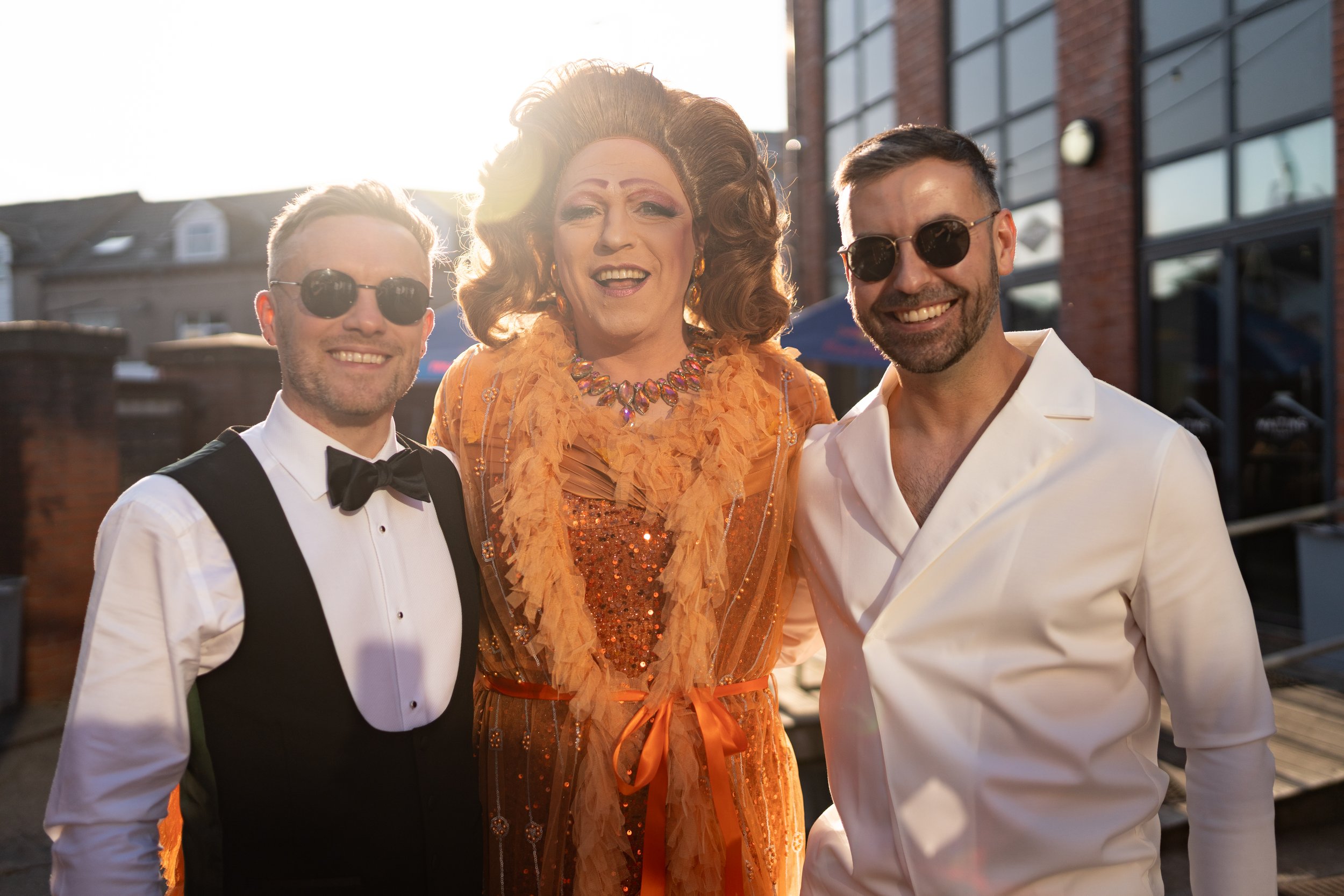 Three people dressed in formal attire smiling outdoors in warm sunlight, with a brick building and outdoor seating in the background.