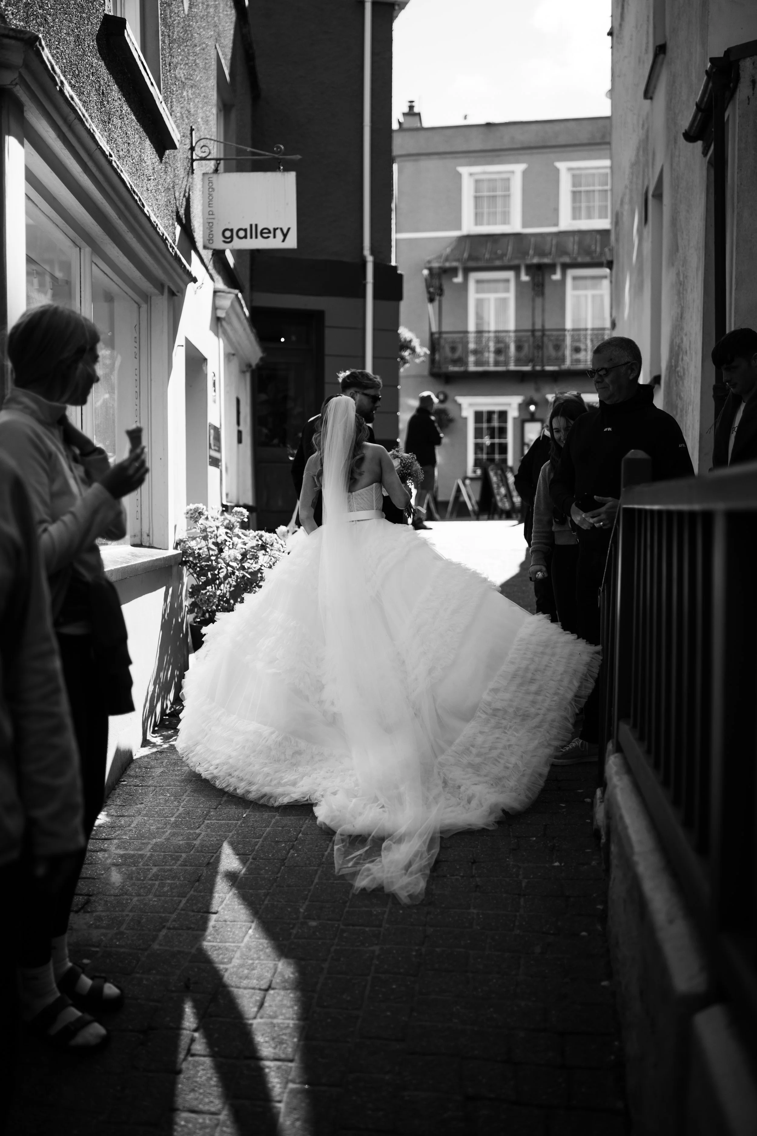 A bride in a wedding dress walking through a narrow street, surrounded by onlookers, with sunlight casting shadows on the ground.