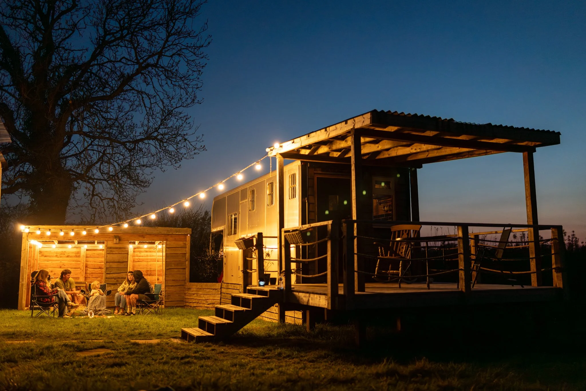 Group of people sitting around a campfire under string lights at dusk, with a trailer and wooden deck in the background.