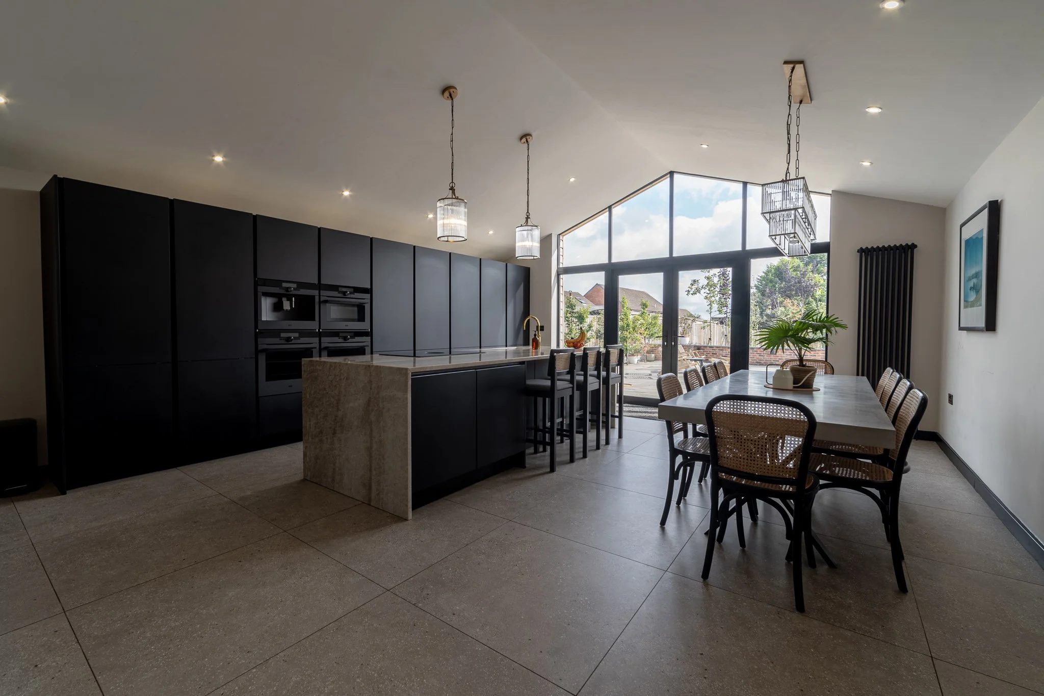 Modern kitchen with black cabinetry, island with marble countertop, dining table with eight chairs, large window with outdoor view, pendant lights, and tiled floor.